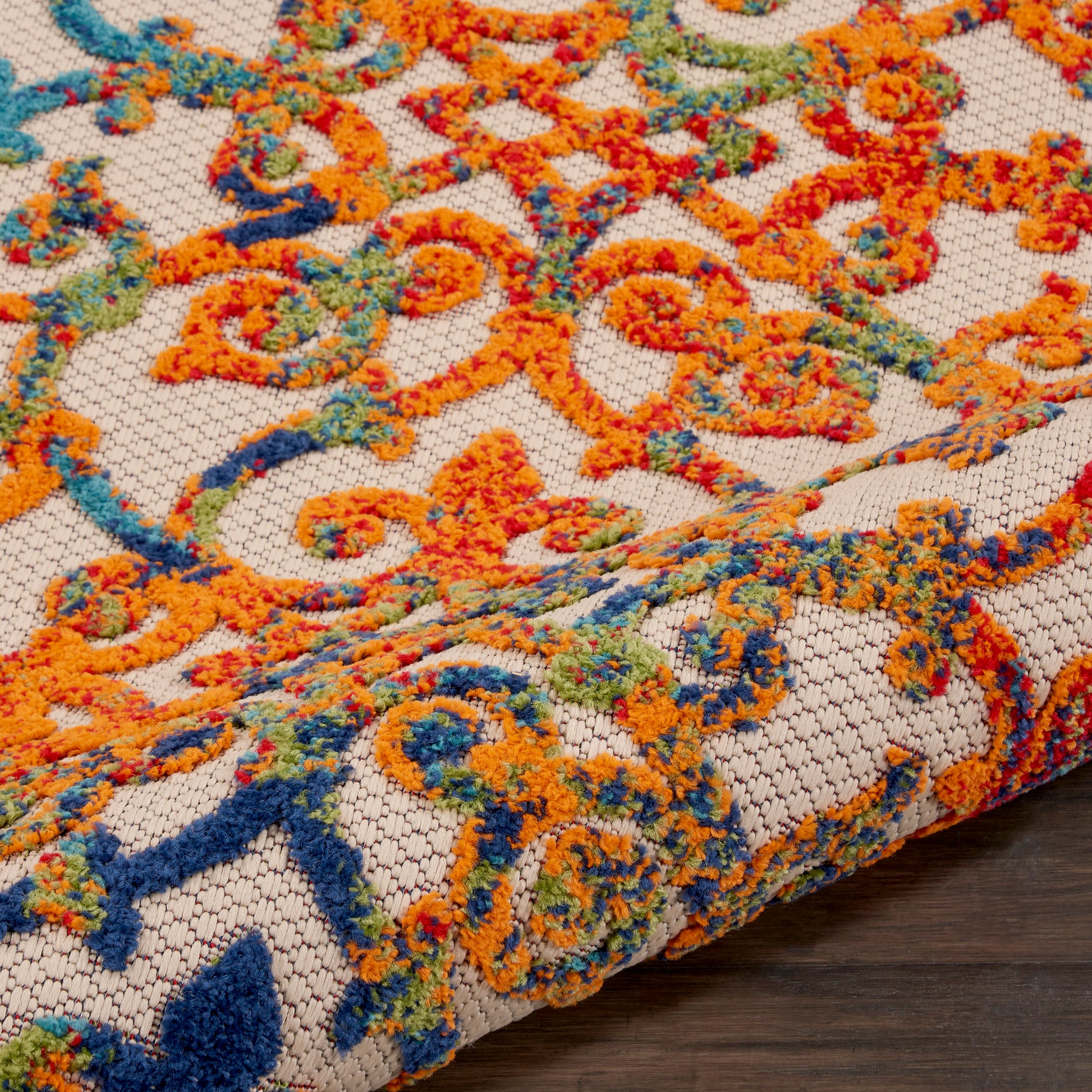 Close-up of a beige outdoor rug with orange, blue, teal & green scrolling floral pattern; looped woven texture.