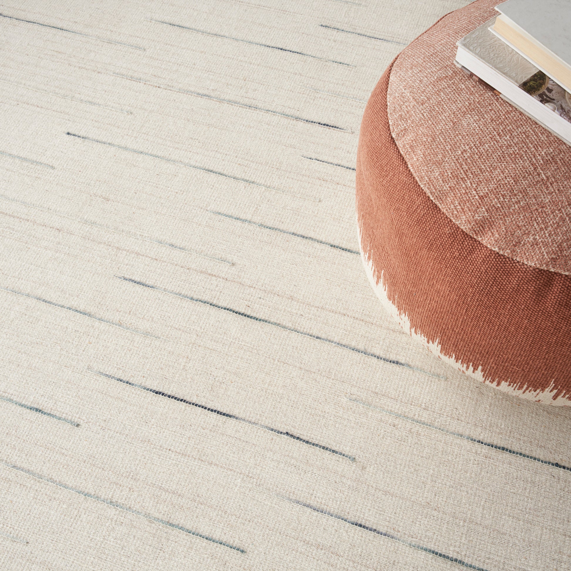Round coral-pink tufted ottoman with fringed edge on a light beige striped rug; a book resting on top.