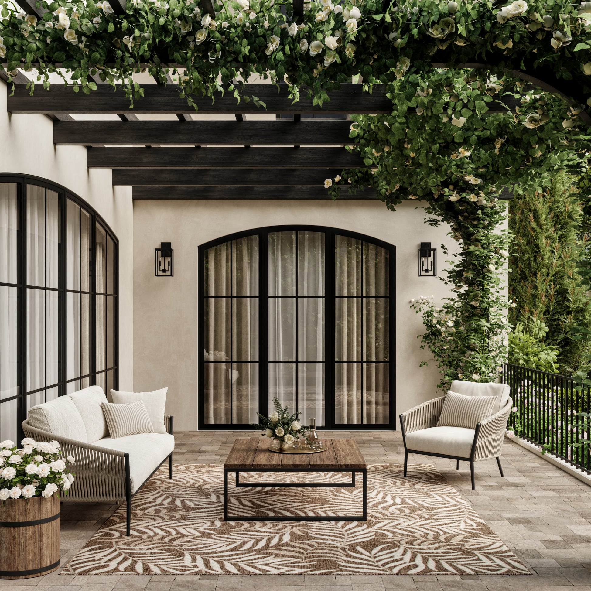 Cozy outdoor lounge under a leafy pergola with arched black-framed doors, cream cushions, and woven rattan seating.