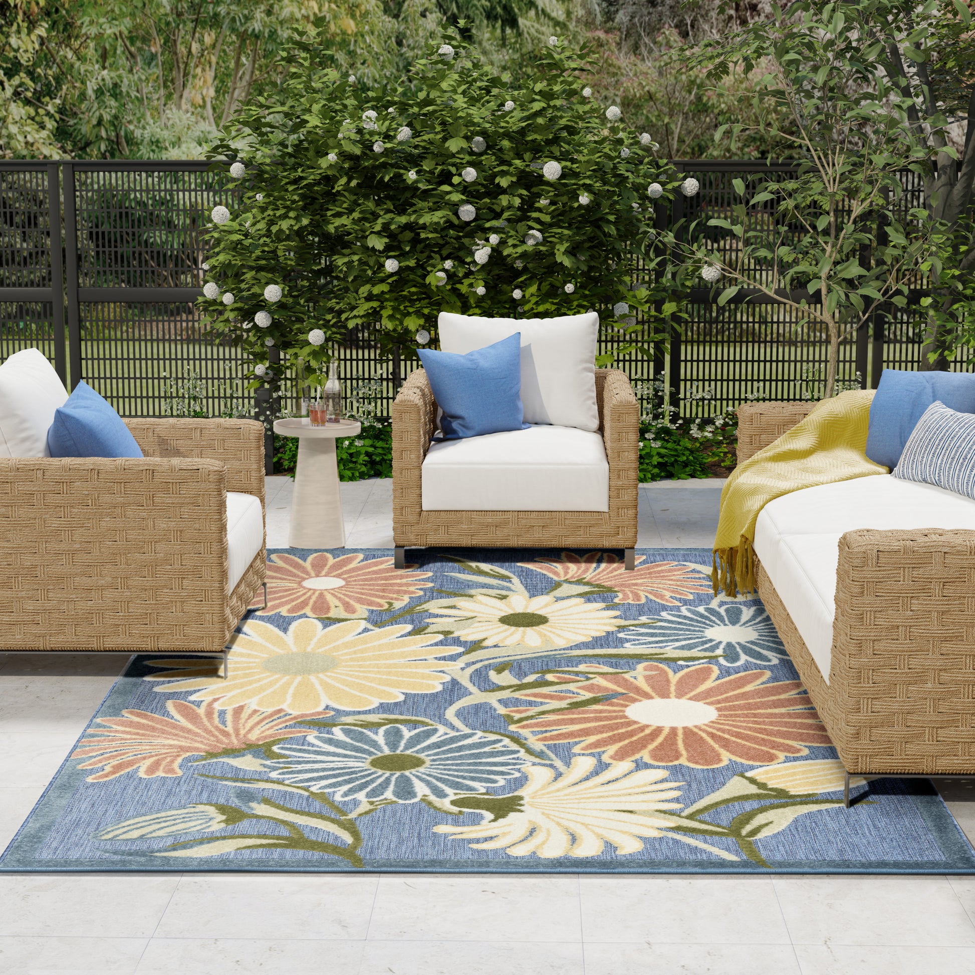Outdoor seating area with wicker furniture, white cushions, blue accents, and a bold floral rug in blue, yellow, orange.