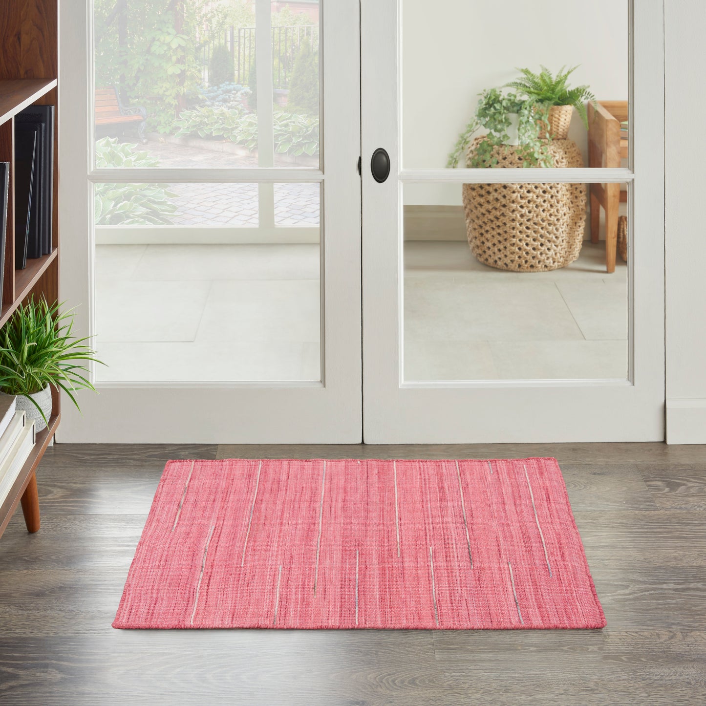 Soft pink rectangular rug on dark wood floor in front of glass doors; greenery and wicker basket in background.