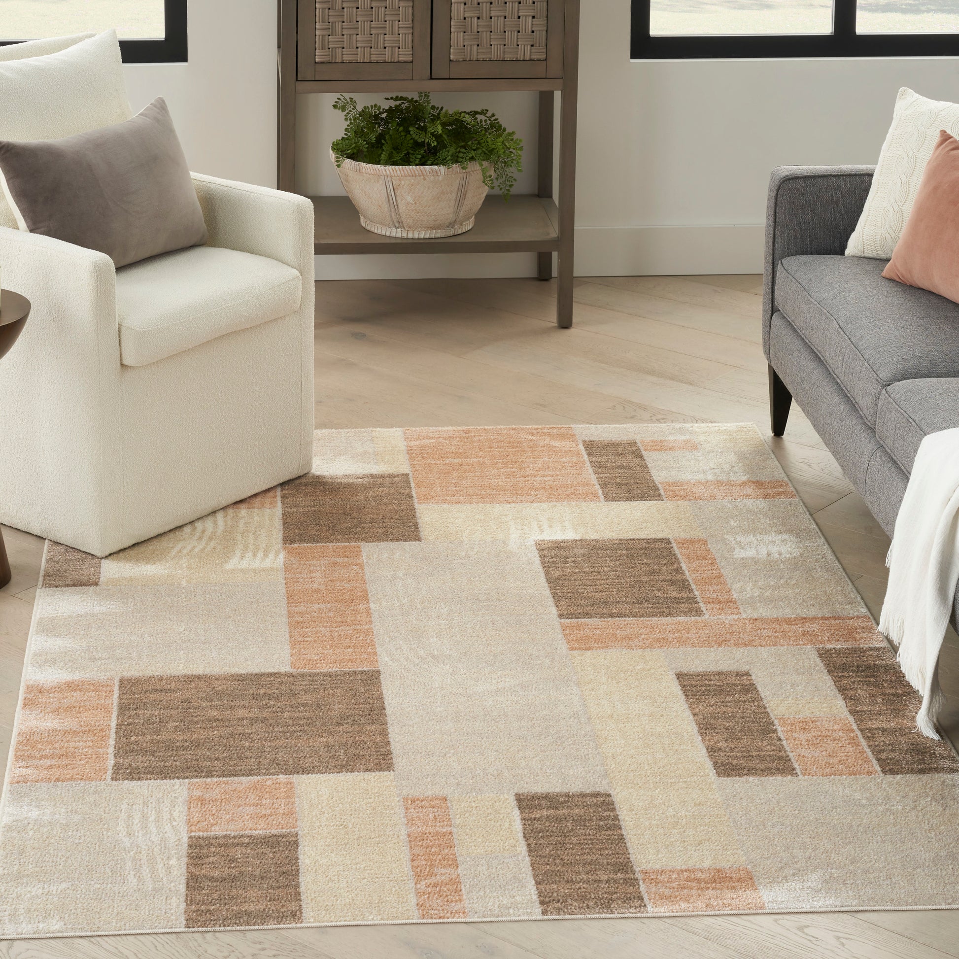 Geometric area rug in beige, tan, peach and brown blocks; modern living room with white chair, gray sofa and potted plant.