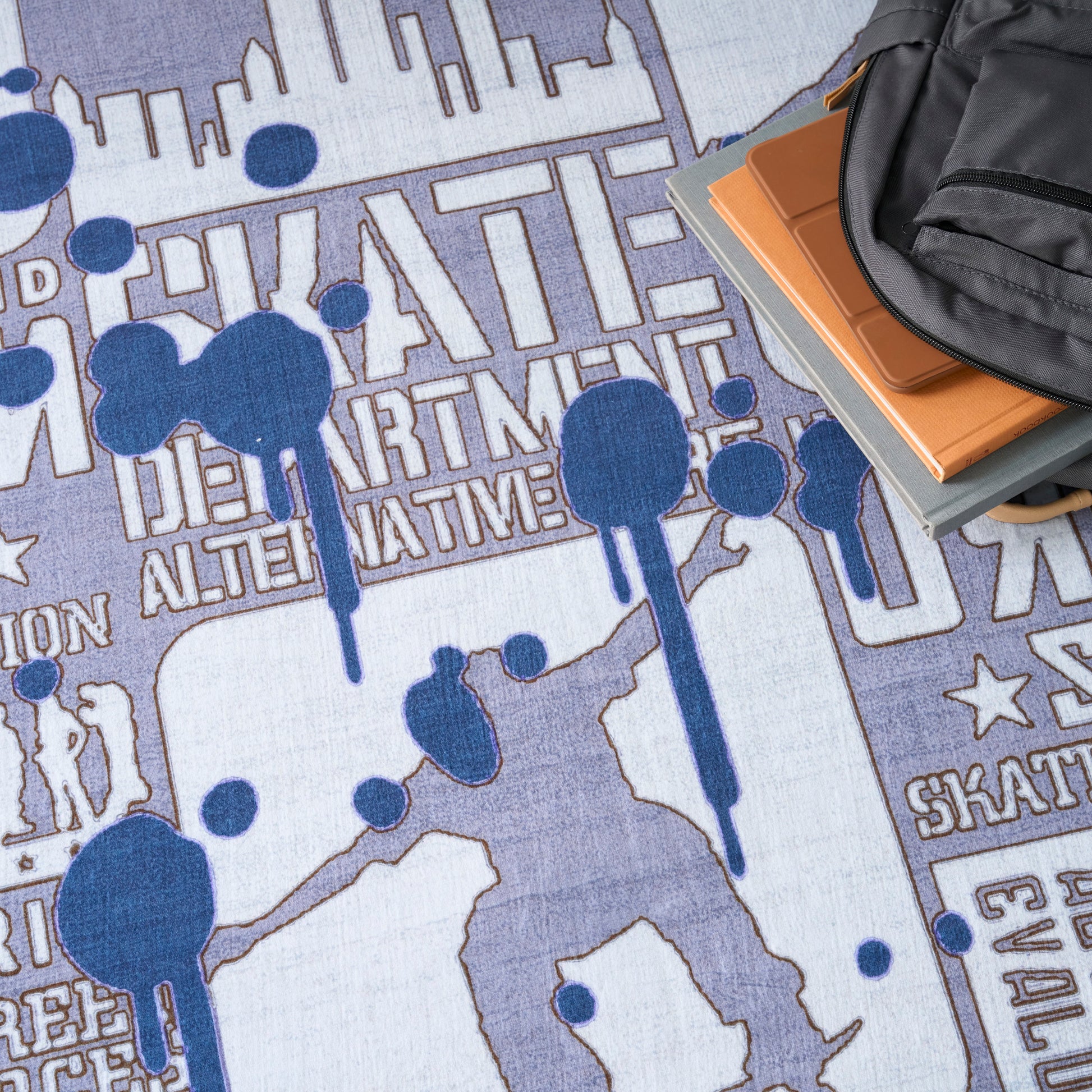 Skate-themed blue and purple rug with skater silhouettes and city skyline; backpack and orange notebook rest on corner.