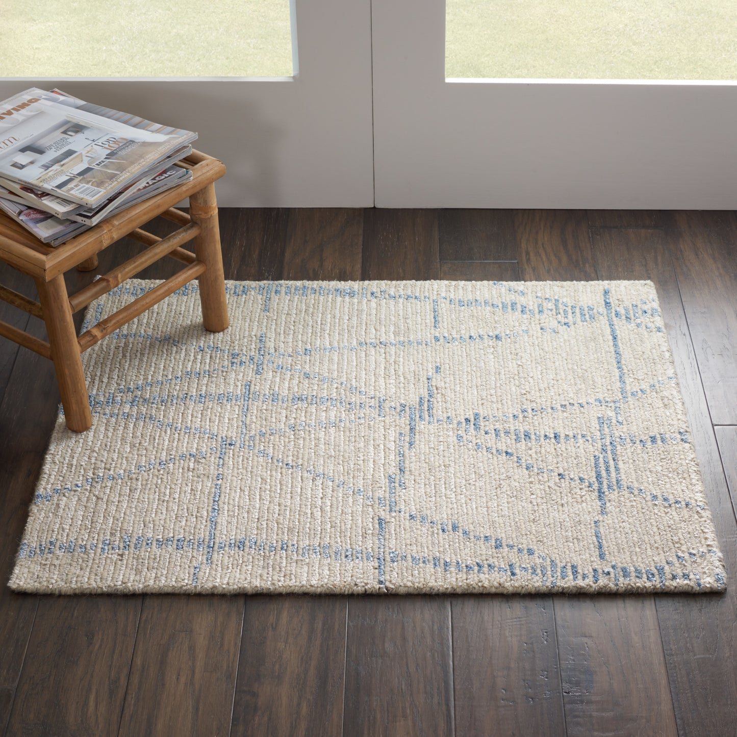 Beige woven area rug with pale blue geometric lines on dark wood floor, beside a small wooden stool with magazines.