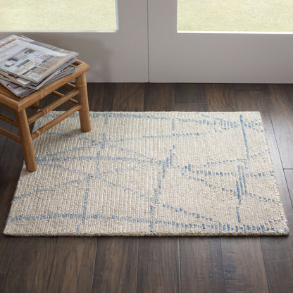 Beige woven area rug with pale blue geometric lines on dark wood floor, beside a small wooden stool with magazines.