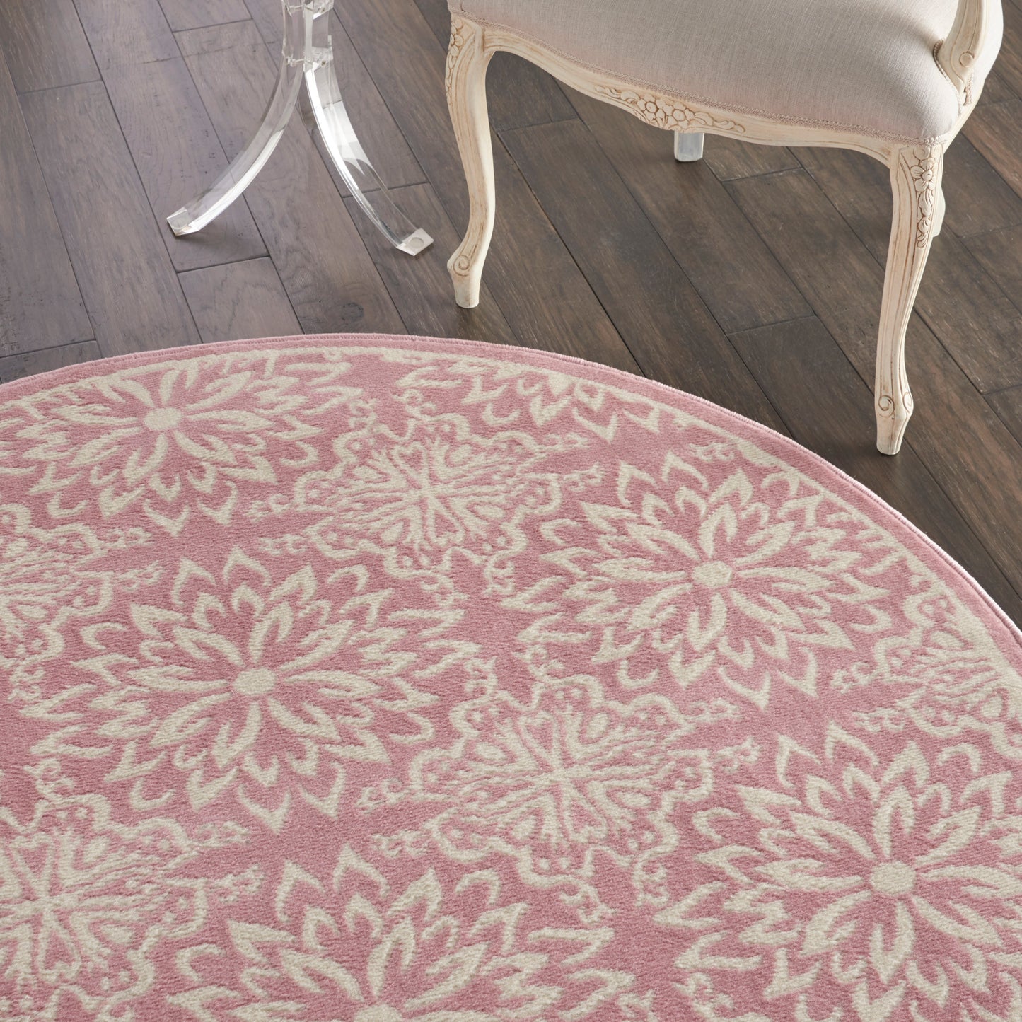 Round pink rug with white floral pattern on dark wood floor; partial cream carved furniture visible.