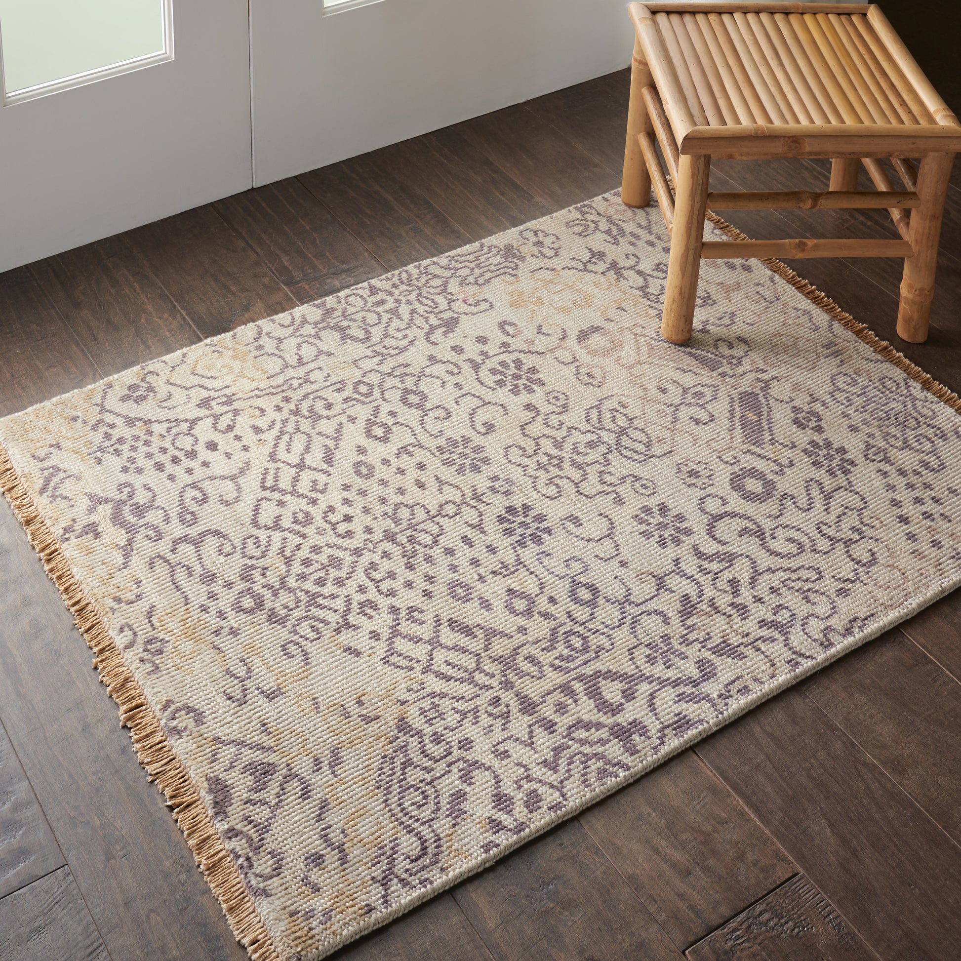 Beige patterned rug with purple floral motifs, fringed edges, laid on dark wood floor beside a bamboo bench.