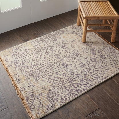 Beige patterned rug with purple floral motifs, fringed edges, laid on dark wood floor beside a bamboo bench.