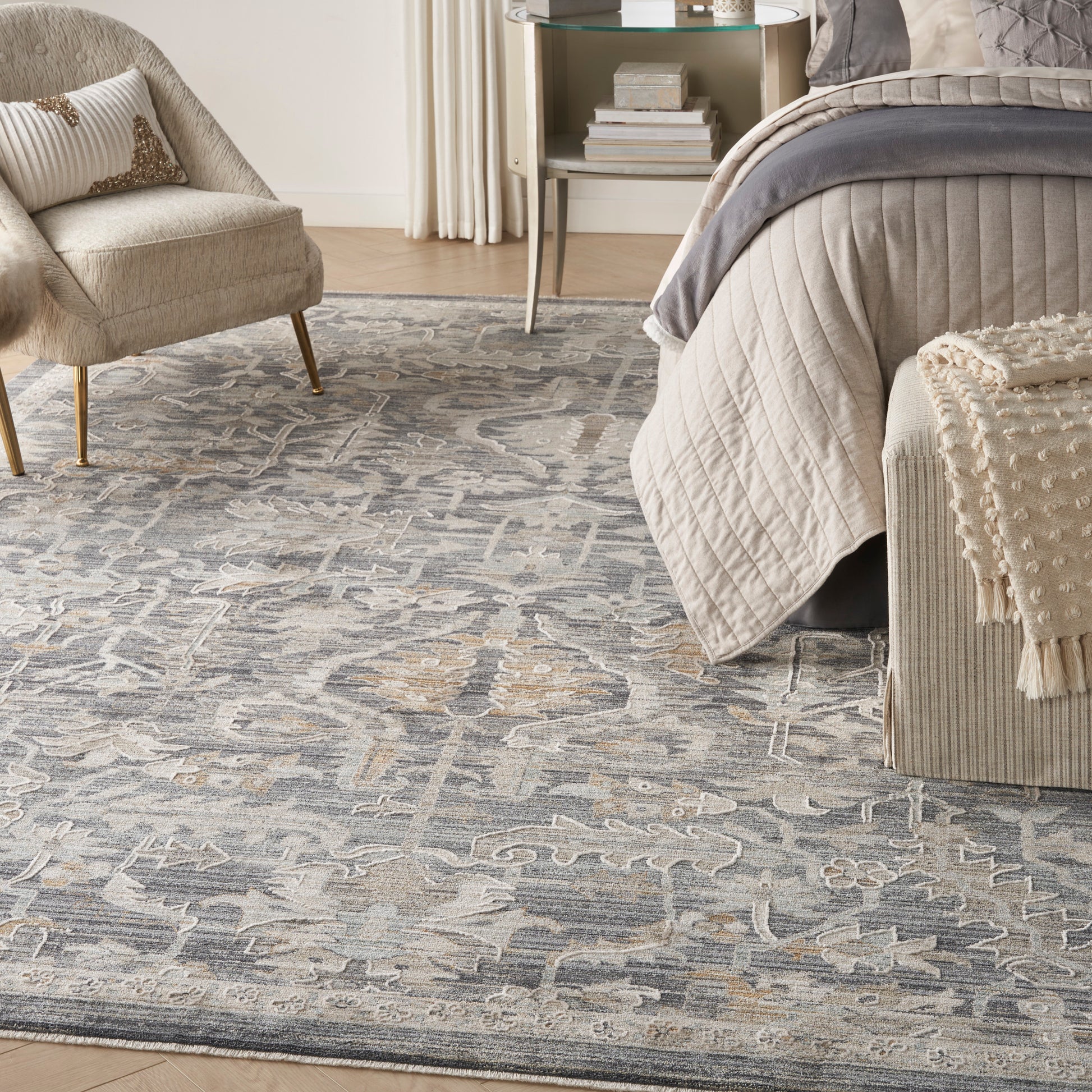 Neutral beige-gray patterned rug in a serene bedroom with a quilted bedspread, cream upholstered chair, glass nightstand, textured throw.