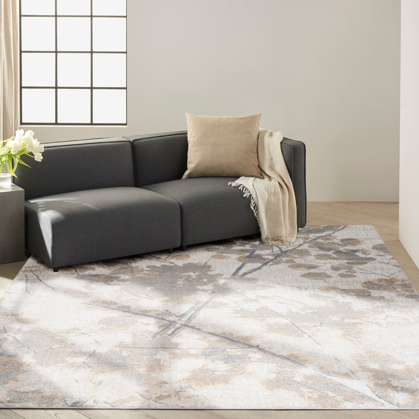 Dark gray fabric sectional with beige cushion and throw on a neutral abstract-pattern rug in a modern living room.