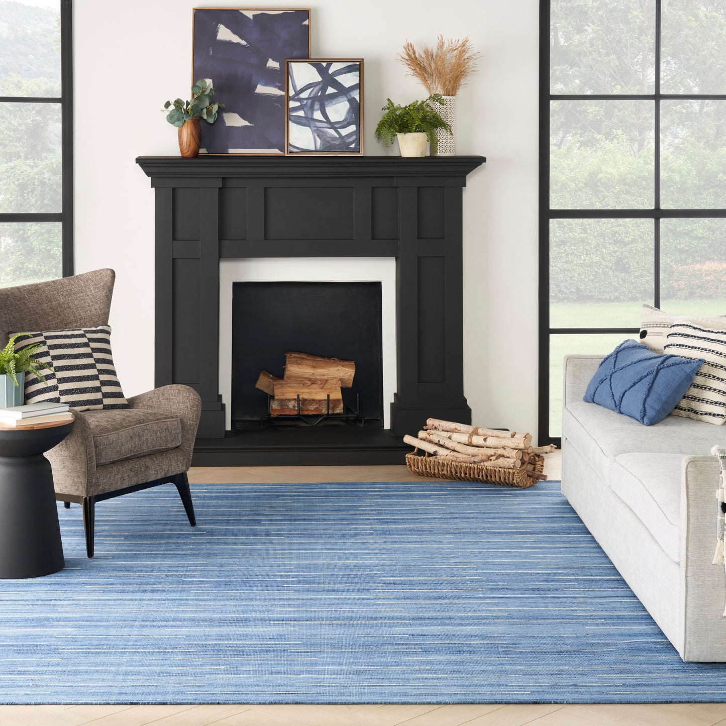 Living room with a matte black fireplace mantel, blue striped rug, gray sofa with pillows, brown armchair & framed art, plants.