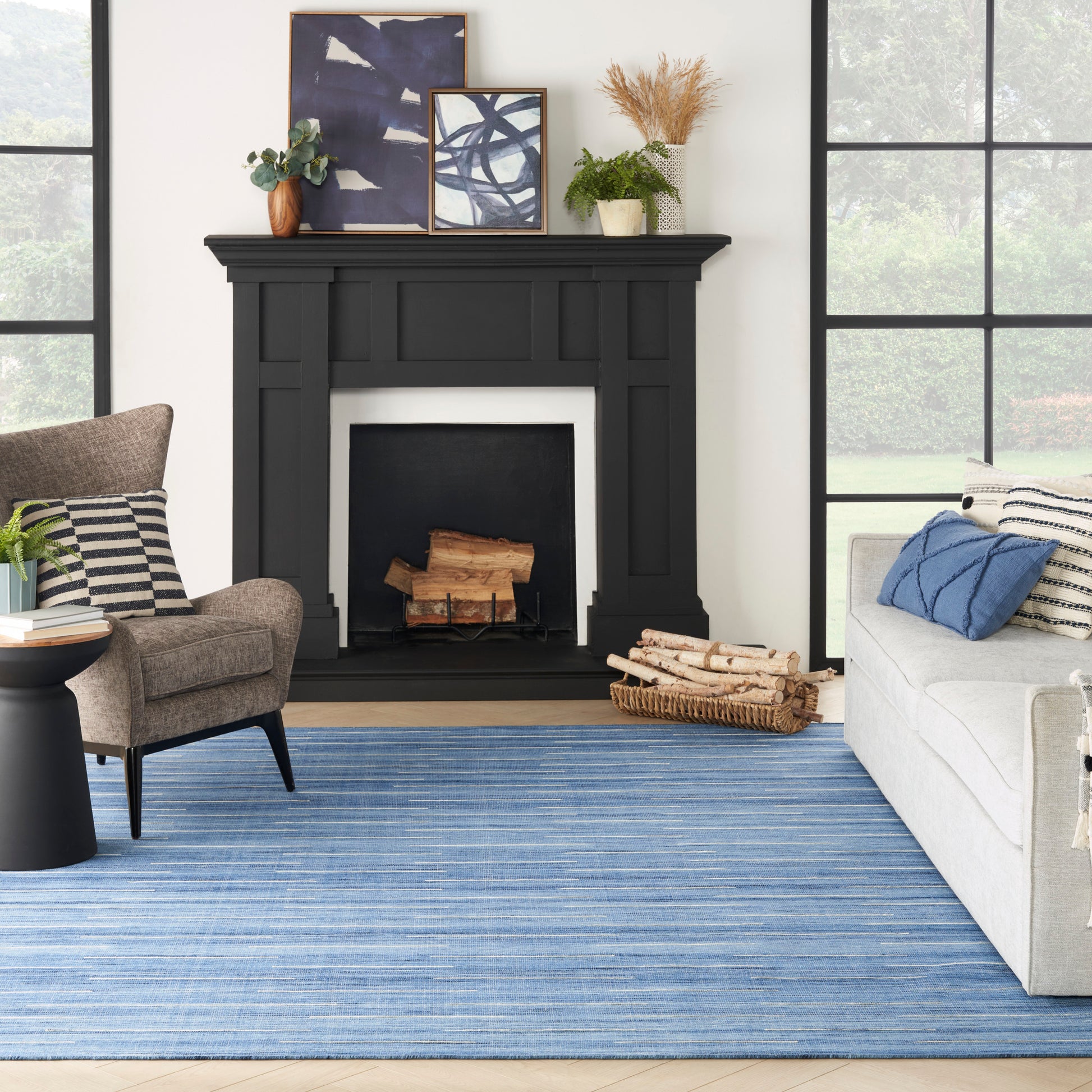 Living room with a matte black fireplace mantel, blue striped rug, gray sofa with pillows, brown armchair & framed art, plants.