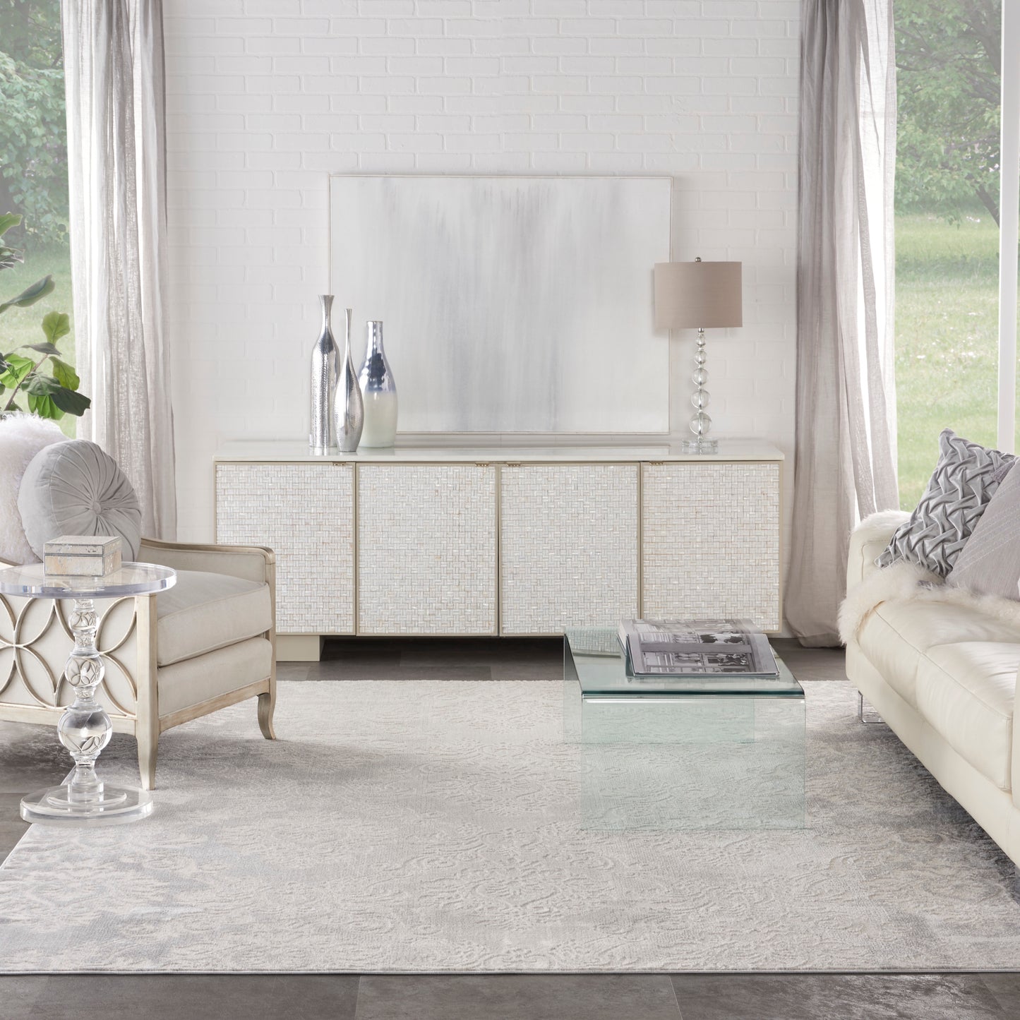 Modern glam living room with white textured sideboard, abstract art, glass coffee table, cream sofas, silver vases, and light gray rug.
