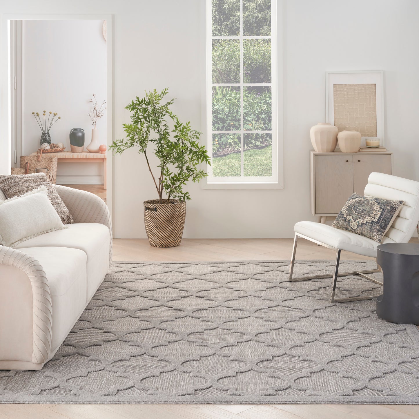 Neutral living room with white sofa, lounge chair, gray geometric rug, tall potted plant, and pale wood accents.