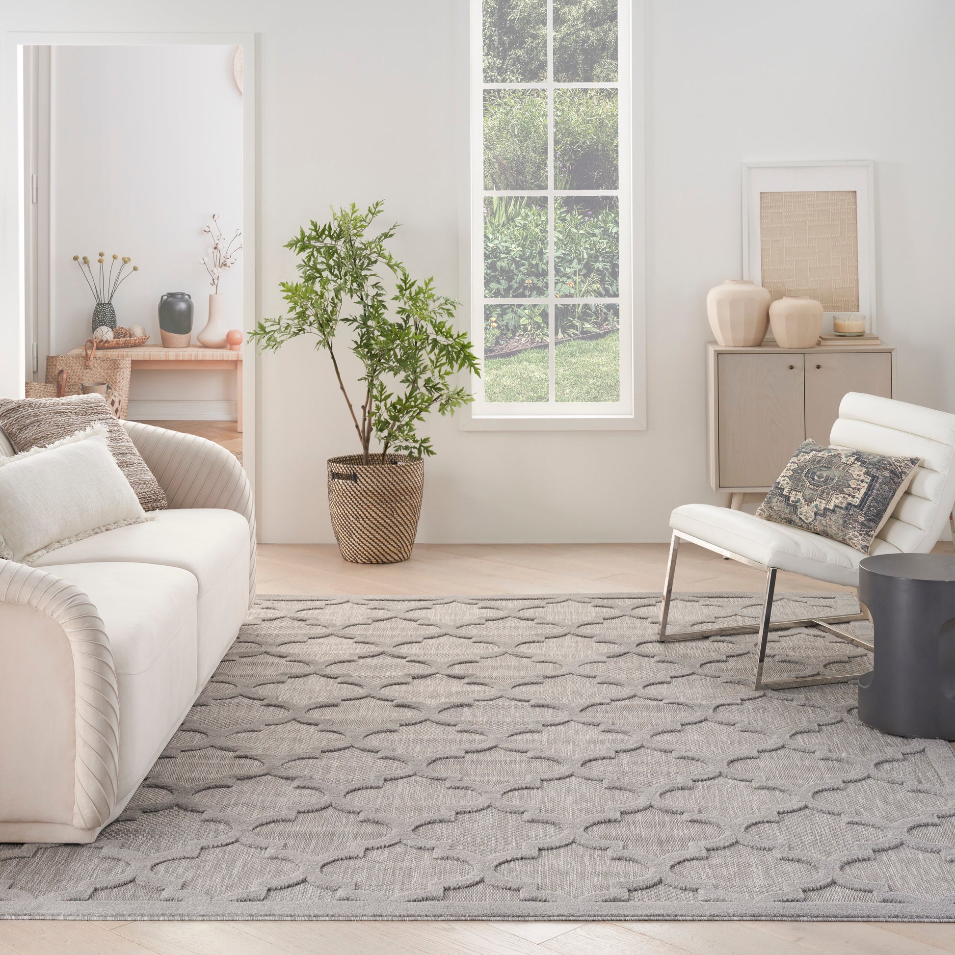 Neutral living room with white sofa, lounge chair, gray geometric rug, tall potted plant, and pale wood accents.