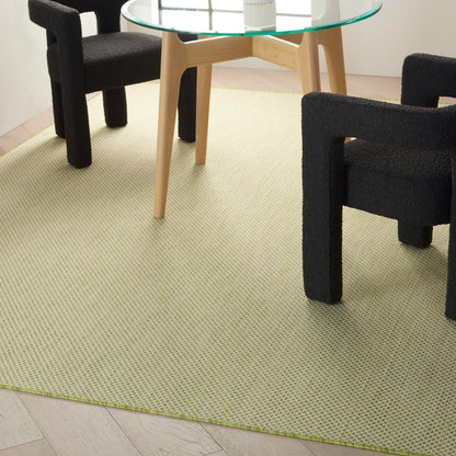 Round glass-top dining table with light wood legs, surrounded by black upholstered block chairs on a pale green woven rug.