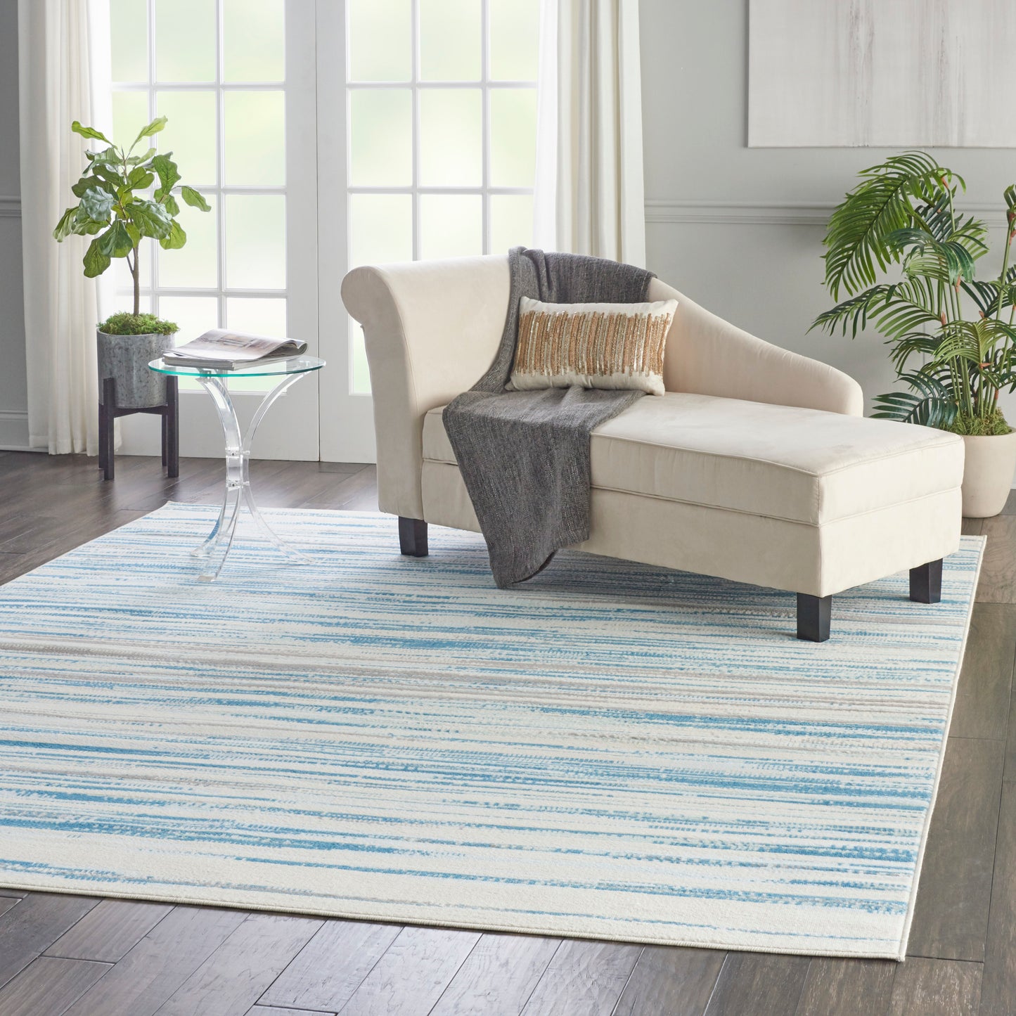 Blue striped rug in a living room scene with a cream chaise lounge, gray throw, gold pillow, glass side table and potted plants.