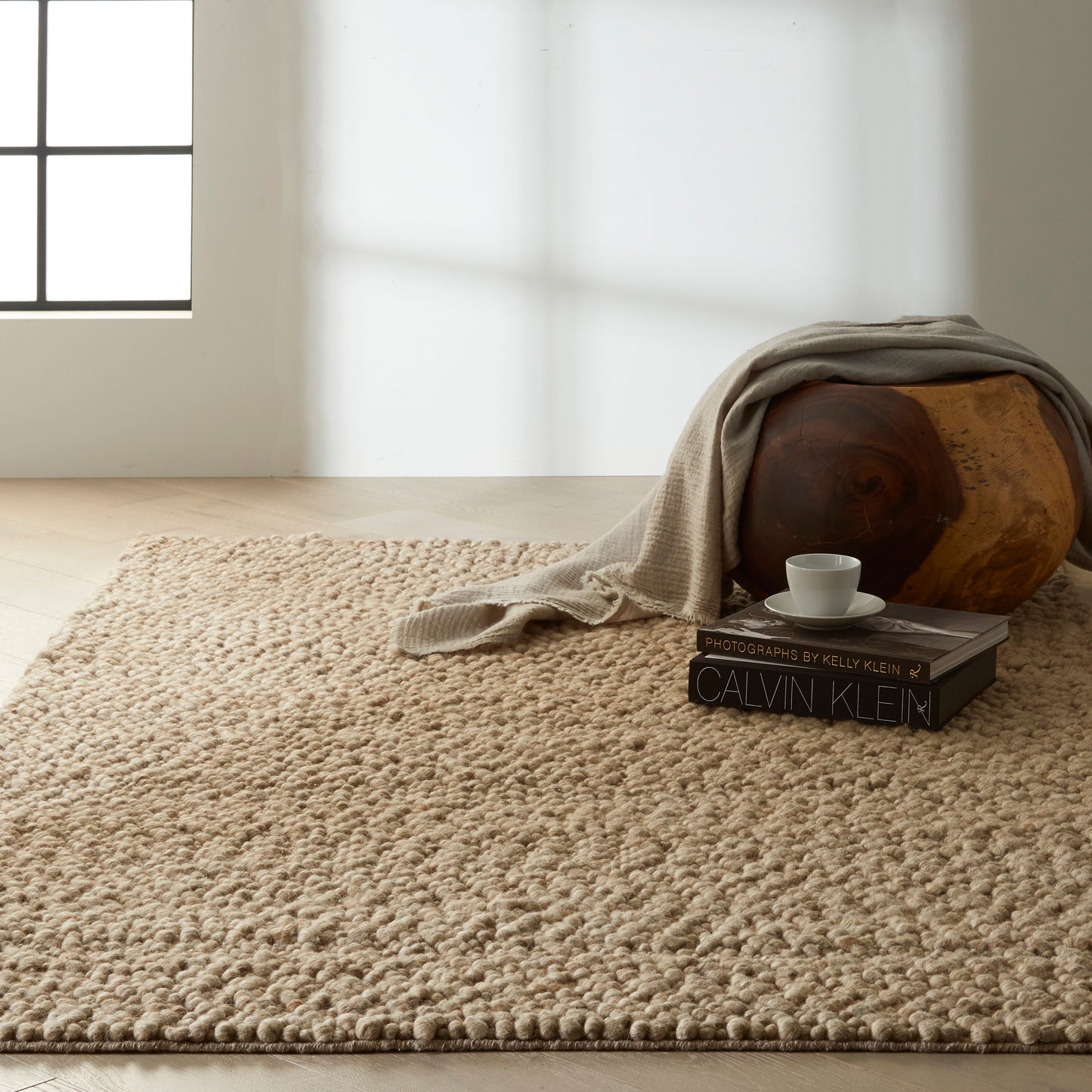 Sunlit living room with beige shag rug, a wooden round piece with a draped linen throw, Calvin Klein books and a cup.