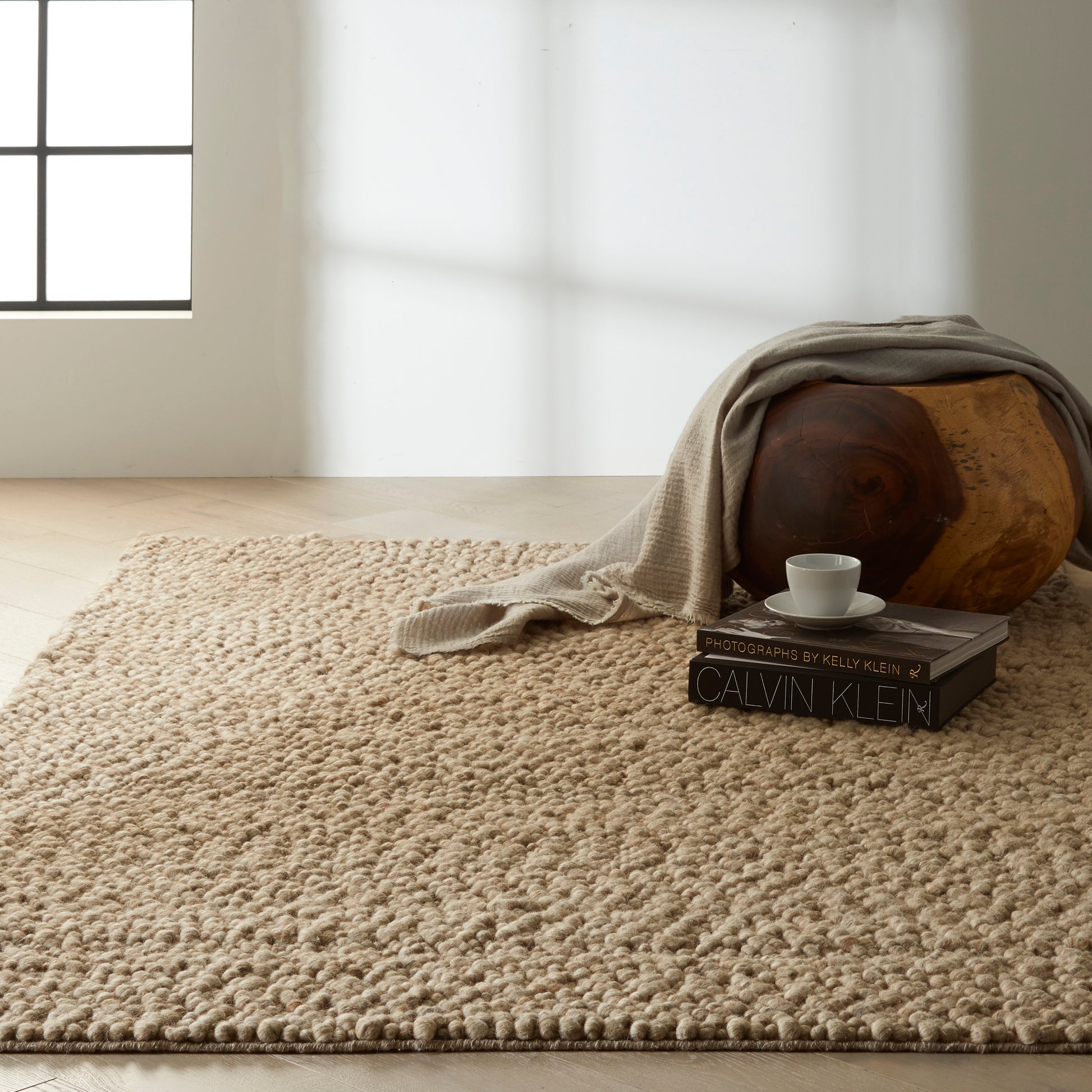 Sunlit living room with beige shag rug, a wooden round piece with a draped linen throw, Calvin Klein books and a cup.