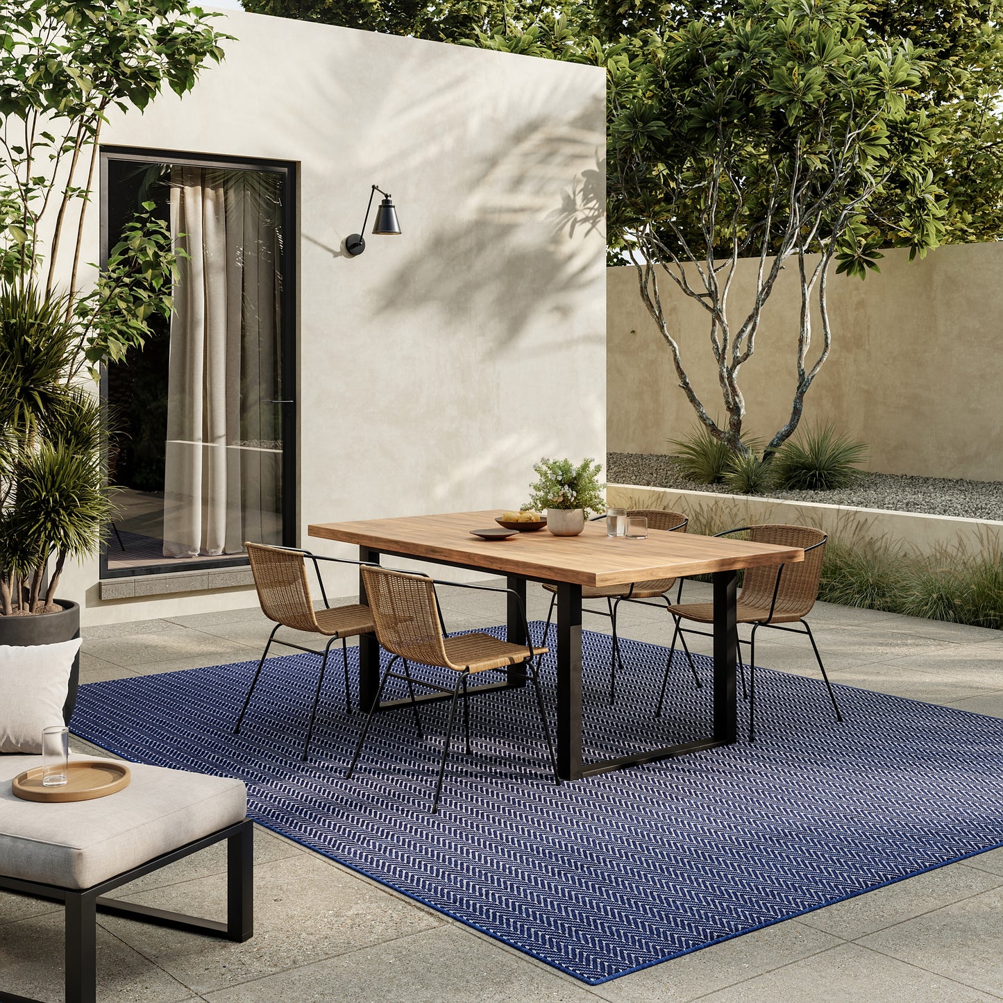 Rectangular wooden dining table with black legs and wicker chairs on a blue patterned outdoor rug in a modern patio.