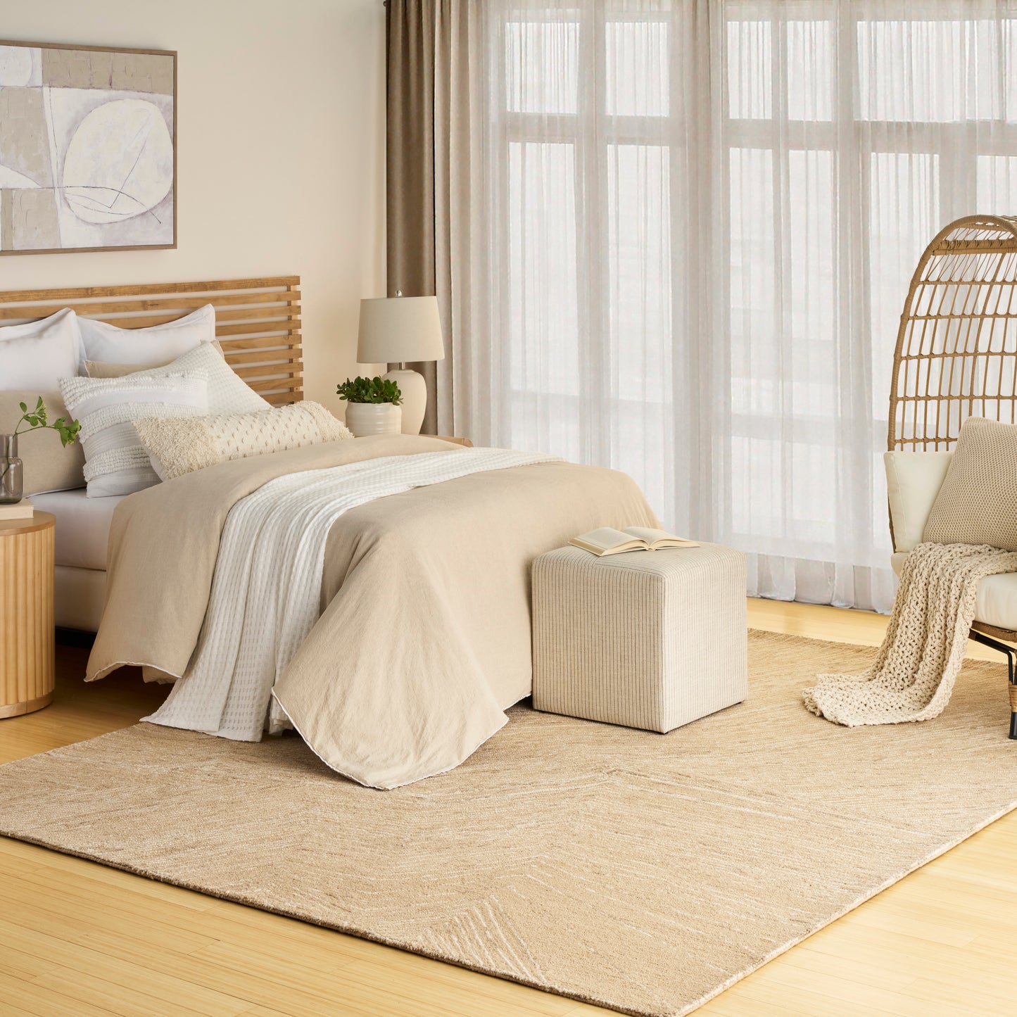 Neutral bedroom scene with a woven rug on the floor; other visible items include a wooden slat bed, beige duvet, white pillows, knit throw, ottoman and wicker chair.