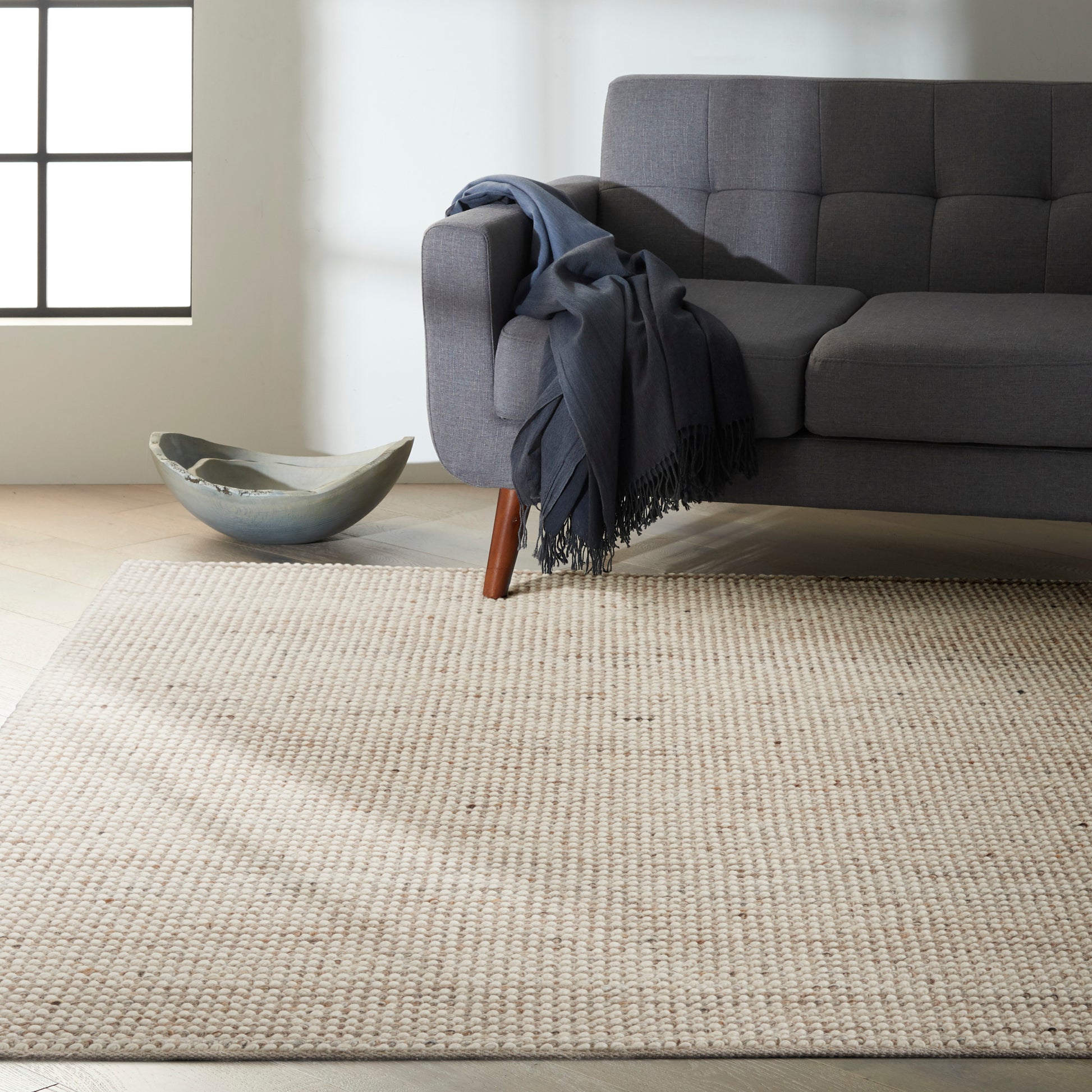 Grey sofa with wooden legs, navy throw on arm, cream woven rug, ceramic bowl on light wood floor by window.