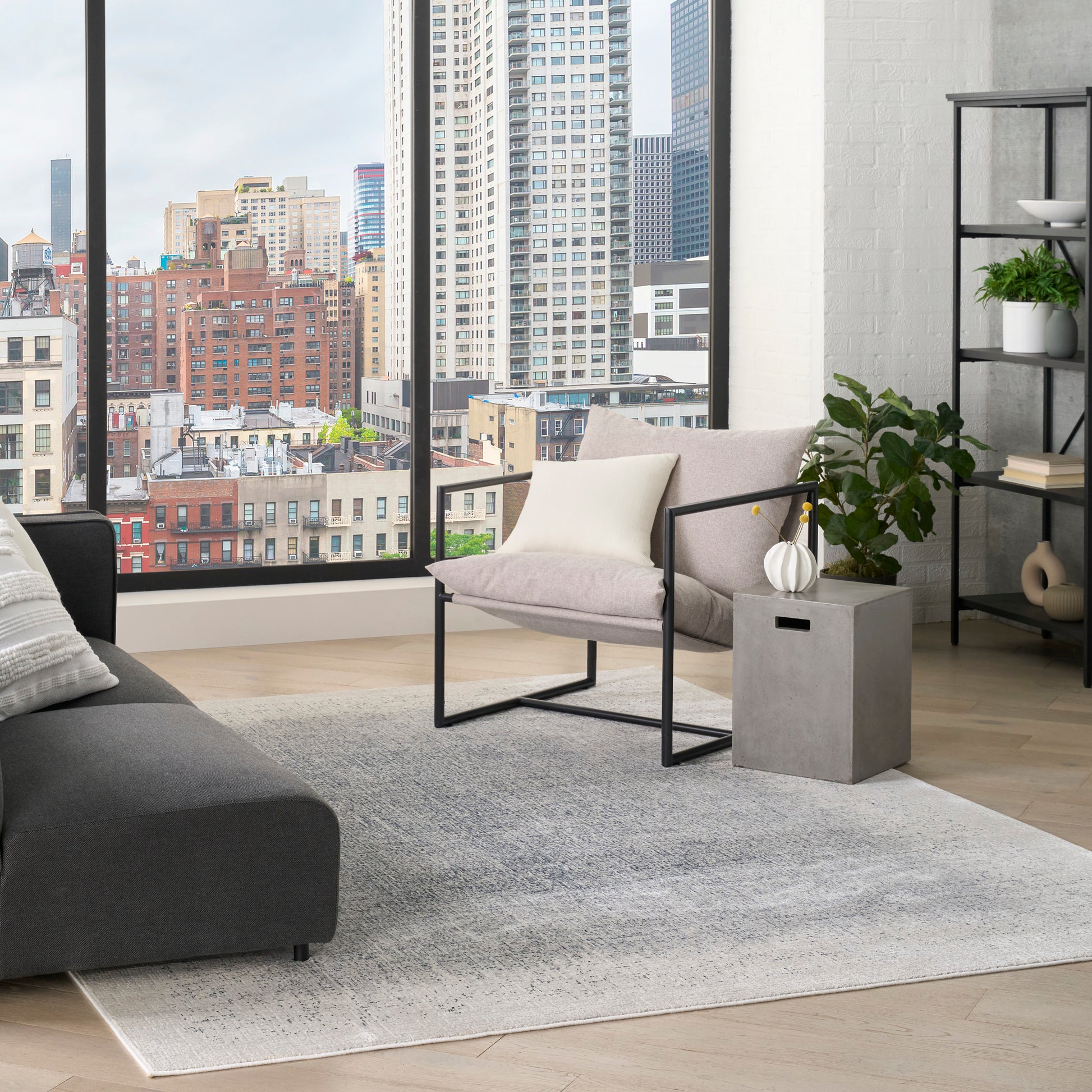 Modern living room with city skyline window, gray sofa, beige metal-framed armchair, gray side table, rug, and shelves.