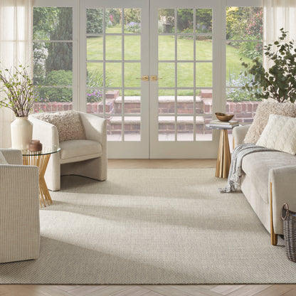 Bright beige living room with French doors, cream sofas and a woven chair, textured rug, gold side tables, greenery outside.