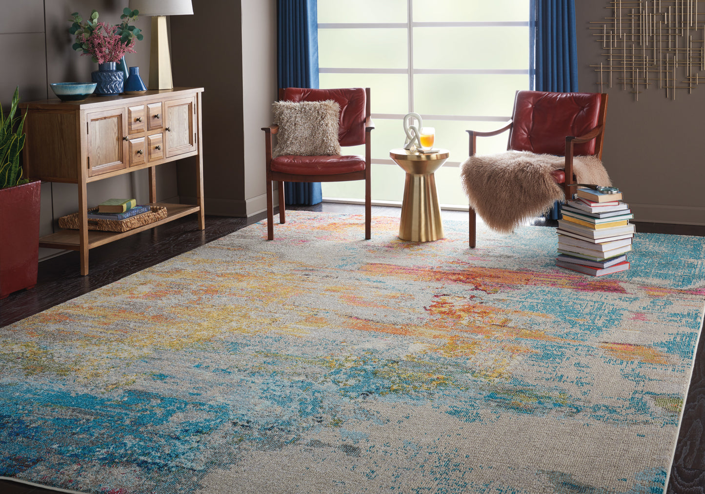 Modern living room with a vibrant abstract multicolor rug, red leather armchairs, fluffy throws, and a gold side table by a sunny window.