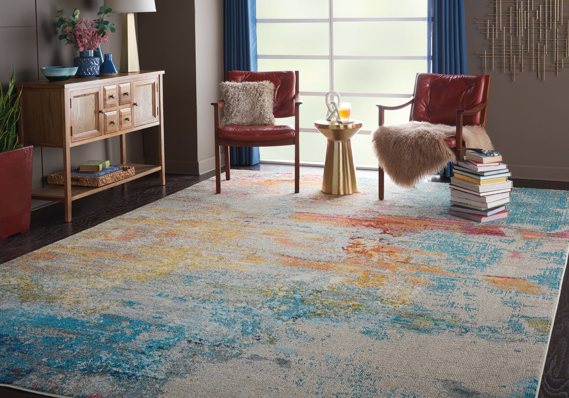 Modern living room with a vibrant abstract multicolor rug, red leather armchairs, fluffy throws, and a gold side table by a sunny window.