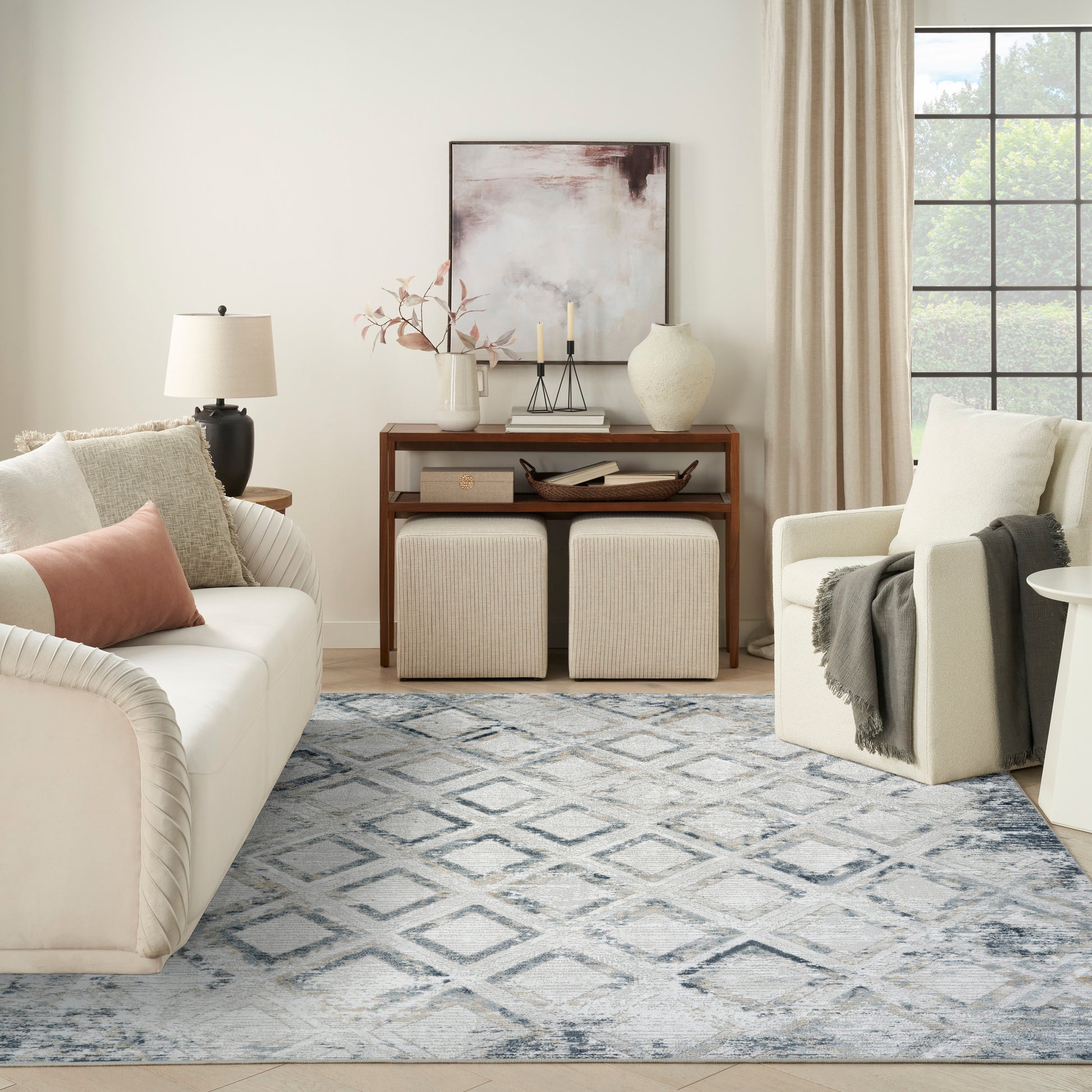 Neutral living room: cream sofa, blush pillow, wooden console, beige ottomans, blue rug, armchair by window.