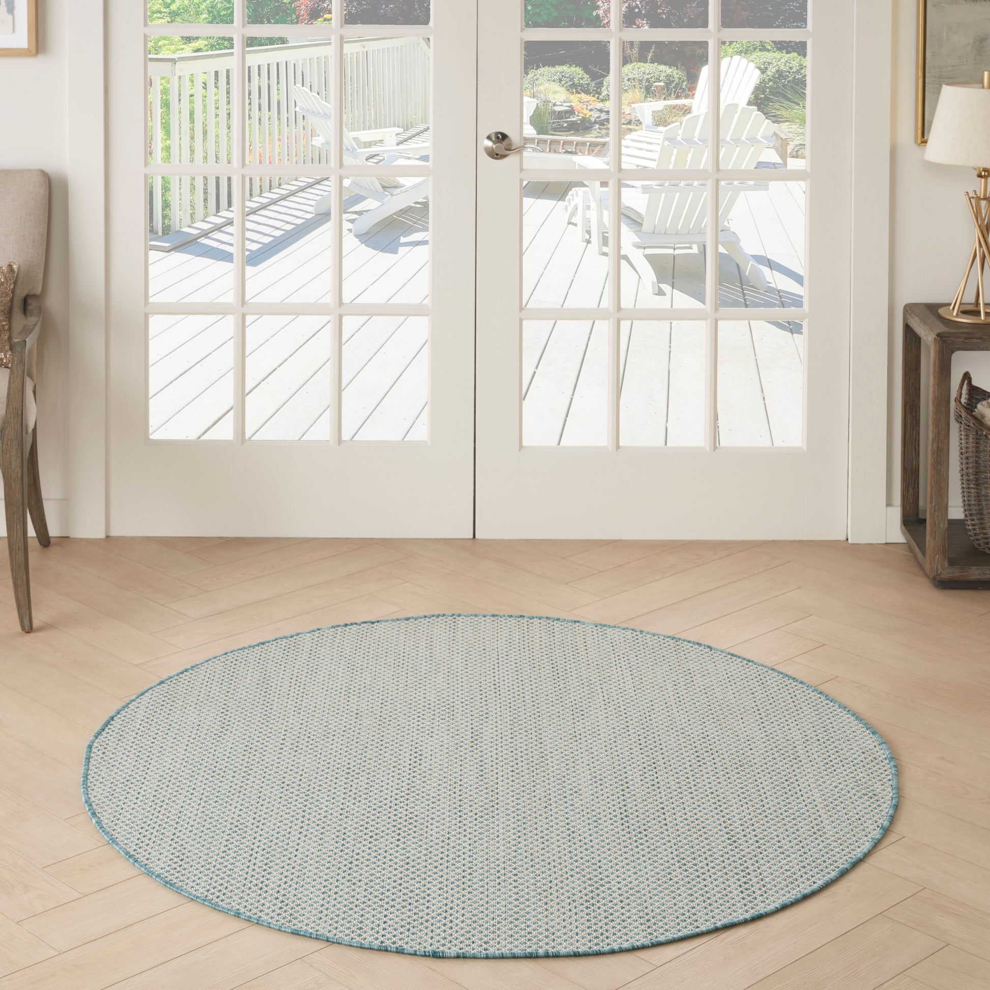 Round blue-gray woven rug with turquoise edge on light wood floor in a bright entry by glass French doors.