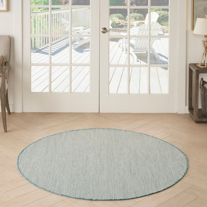 Round blue-gray woven rug with turquoise edge on light wood floor in a bright entry by glass French doors.