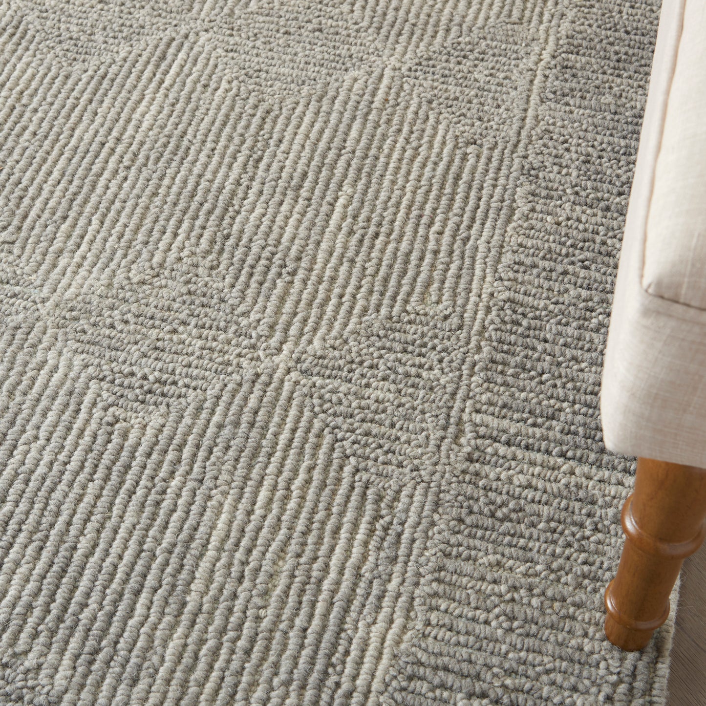 Gray textured area rug with raised loop pattern in circular and linear motifs, next to a light beige upholstered sofa leg.