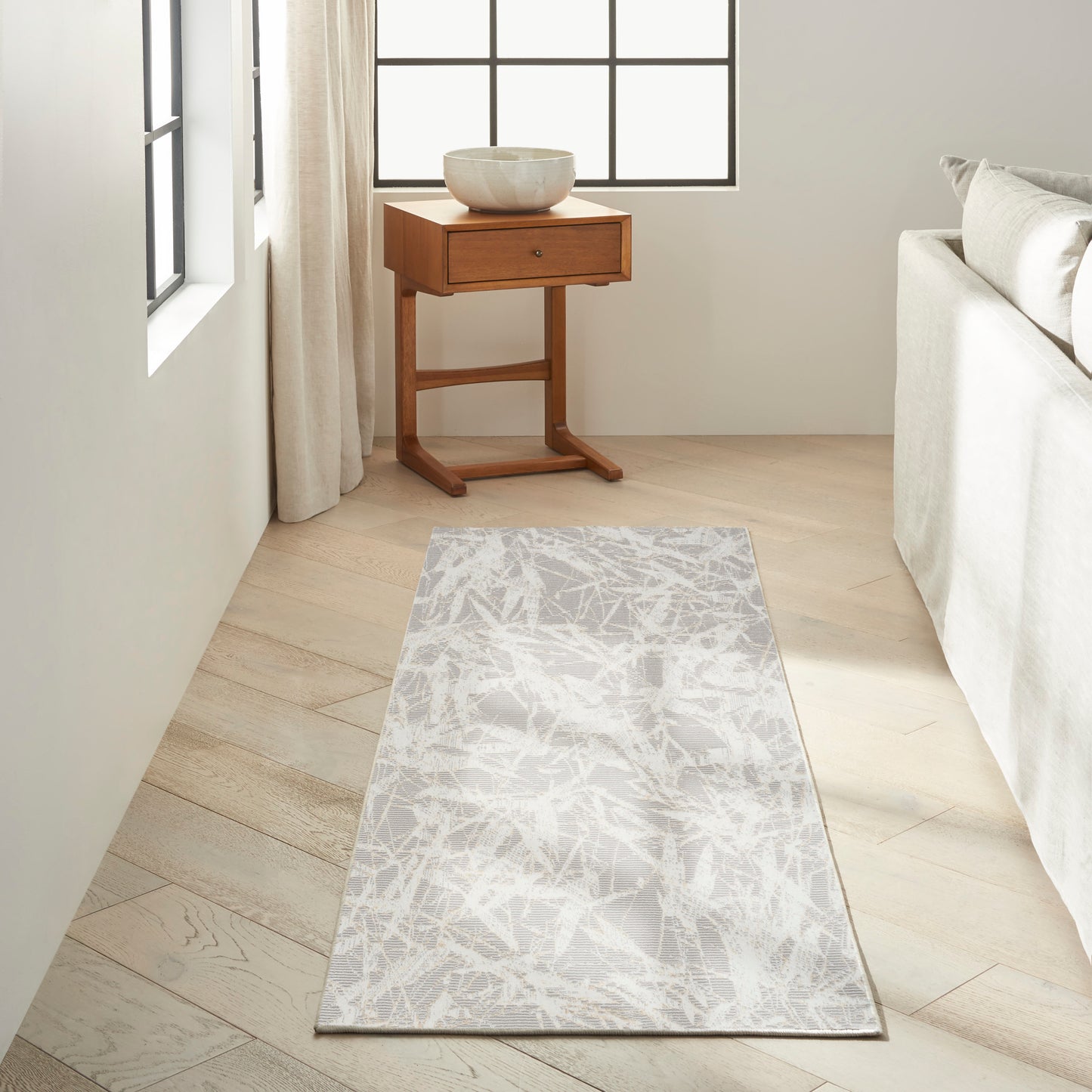 Light-gray patterned runner rug on pale wood floor beside a sofa; small wooden side table with a ceramic bowl near window.
