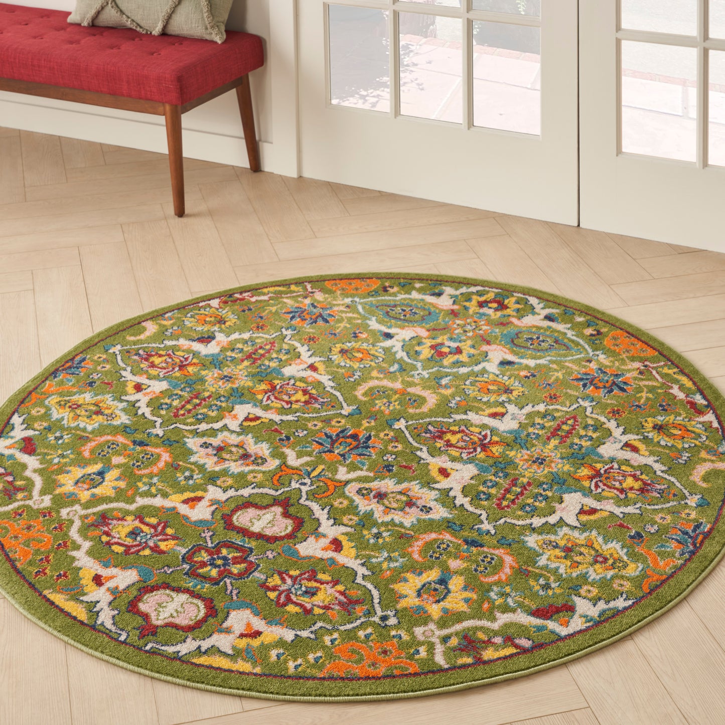Circular green-bordered rug with colorful floral lattice on light wood floor; red cushioned bench by glass doors.