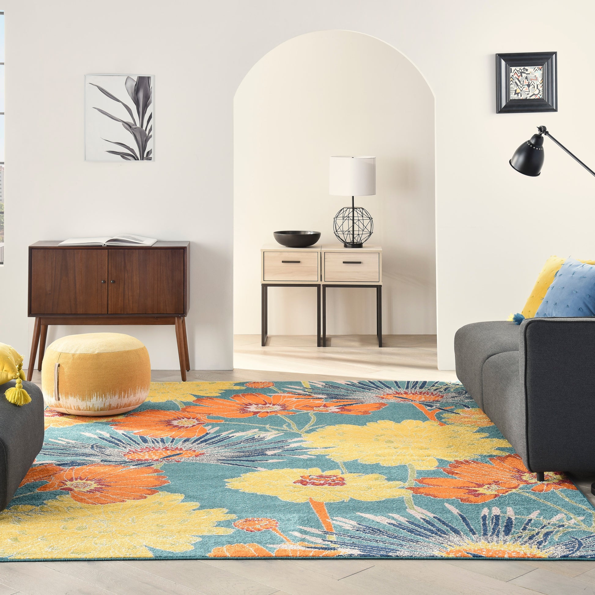 Living room with vibrant floral rug, mid-century furniture, yellow pouf, grey sofa, arched doorway, wood cabinet, floor lamp.
