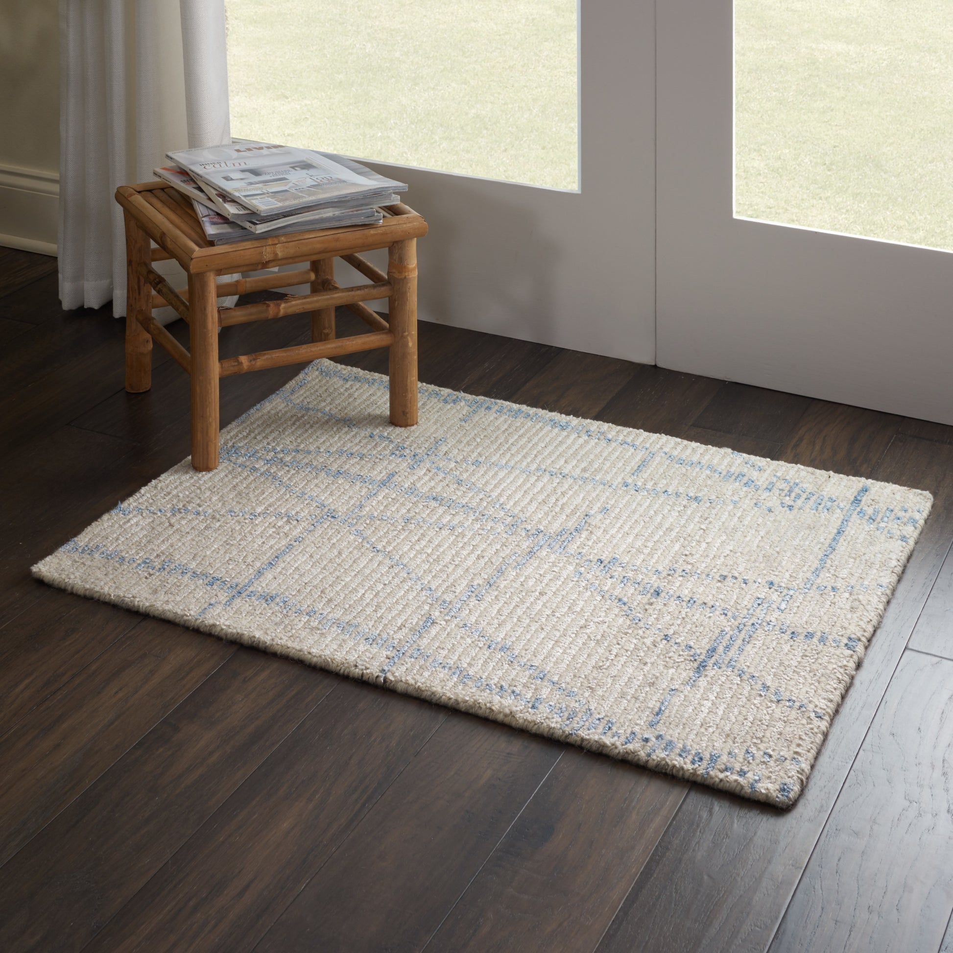 Beige shaggy rug with blue grid pattern on dark hardwood floor; small wooden stool with magazines beside glass doors.