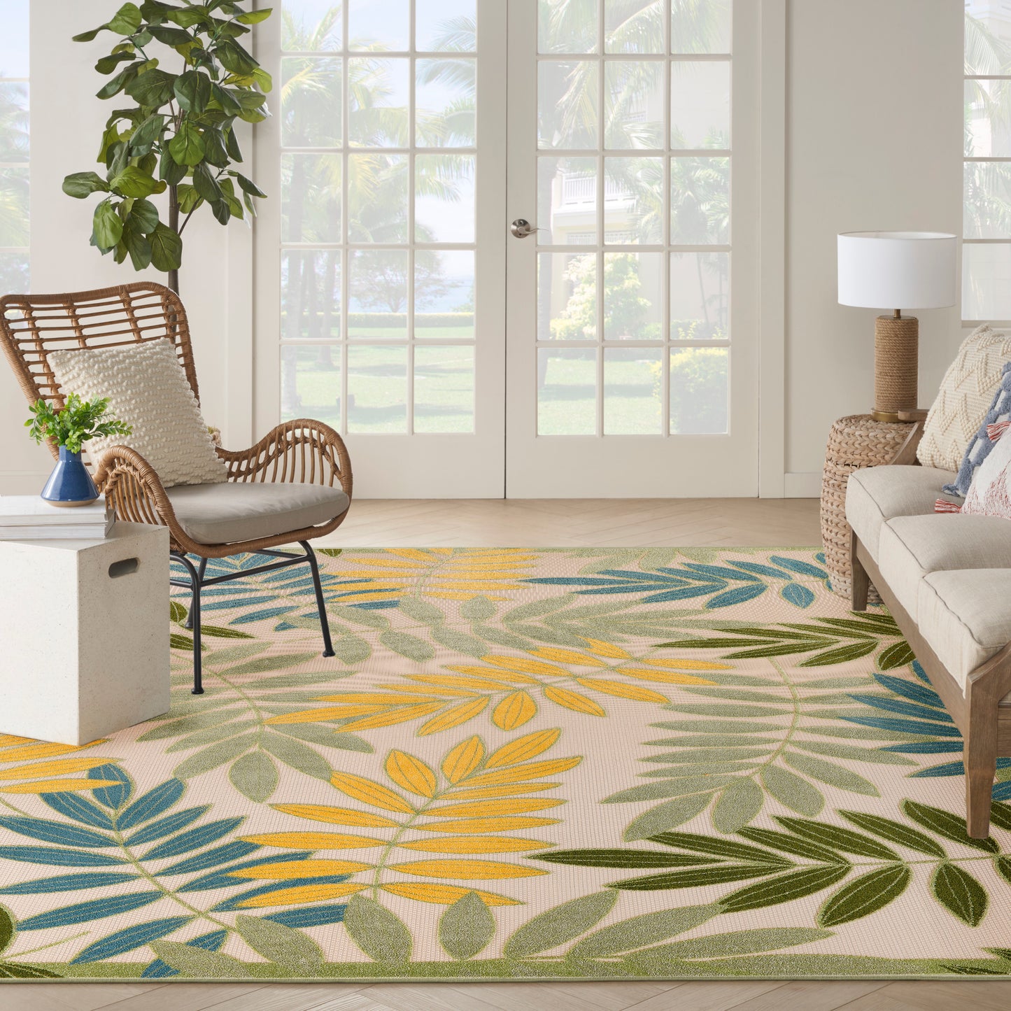 Living room with tropical leaf rug in yellow, blue, green; wicker chair, sofa, plant, and French doors.