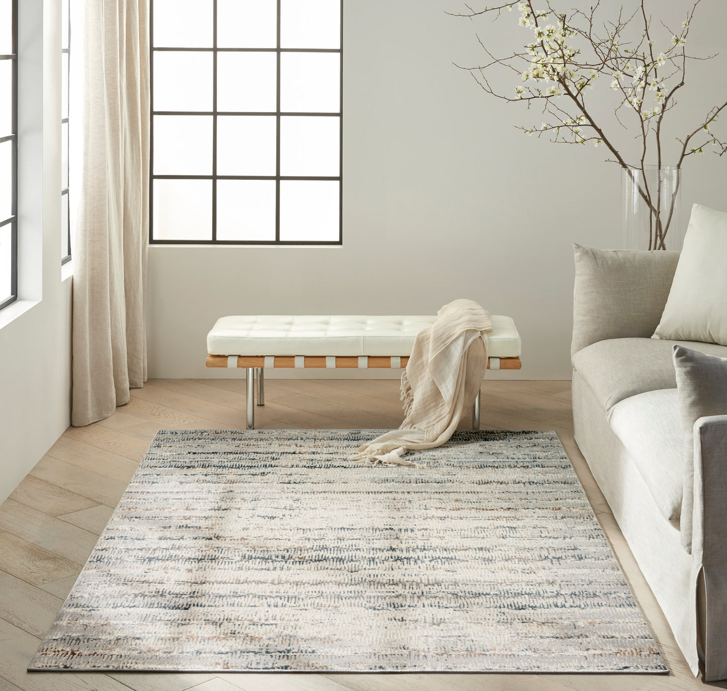 Bright living room with white tufted bench on wood slats, beige throw, gray sofa, blue-gray patterned rug.