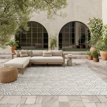 Outdoor patio with beige sectional sofa on light wood frame, patterned gray rug, woven pouf, arched windows, potted trees.