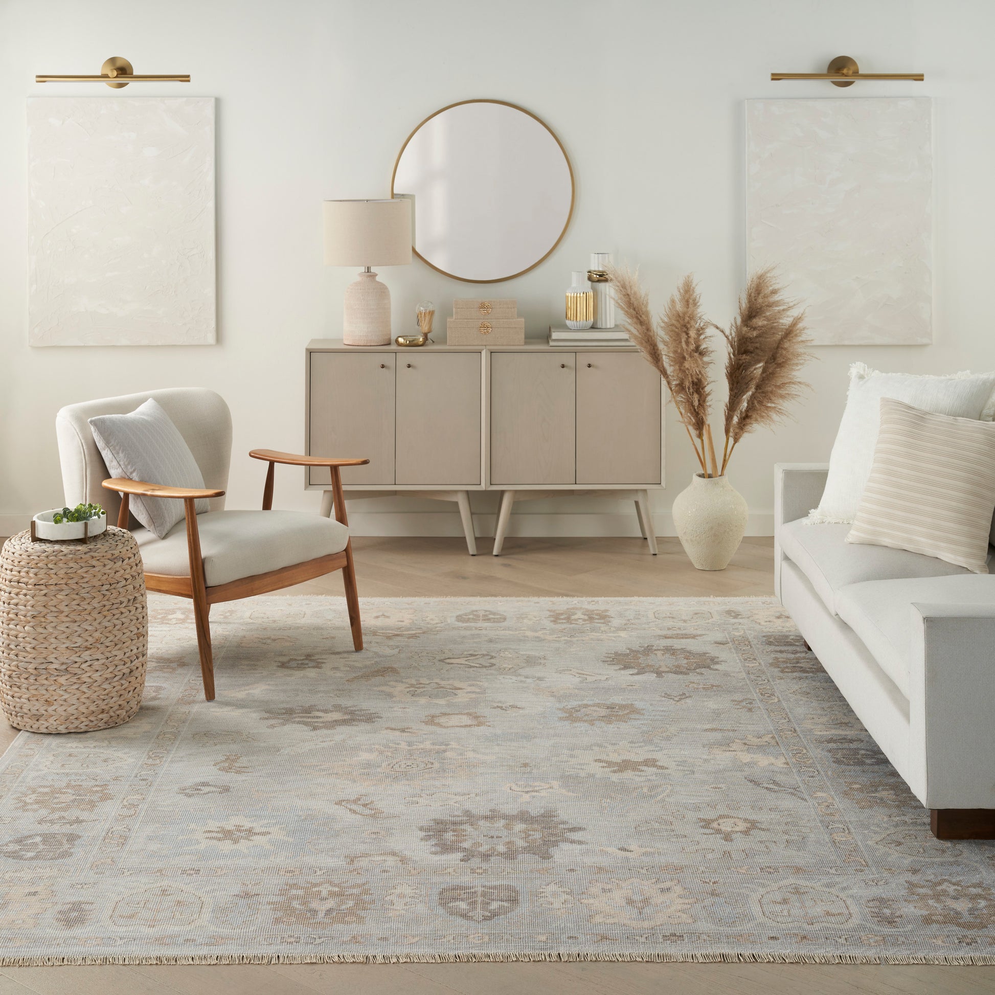 Neutral living room with gray sofa, mid-century chair, woven ottoman, rug, console with round mirror and pampas grass.