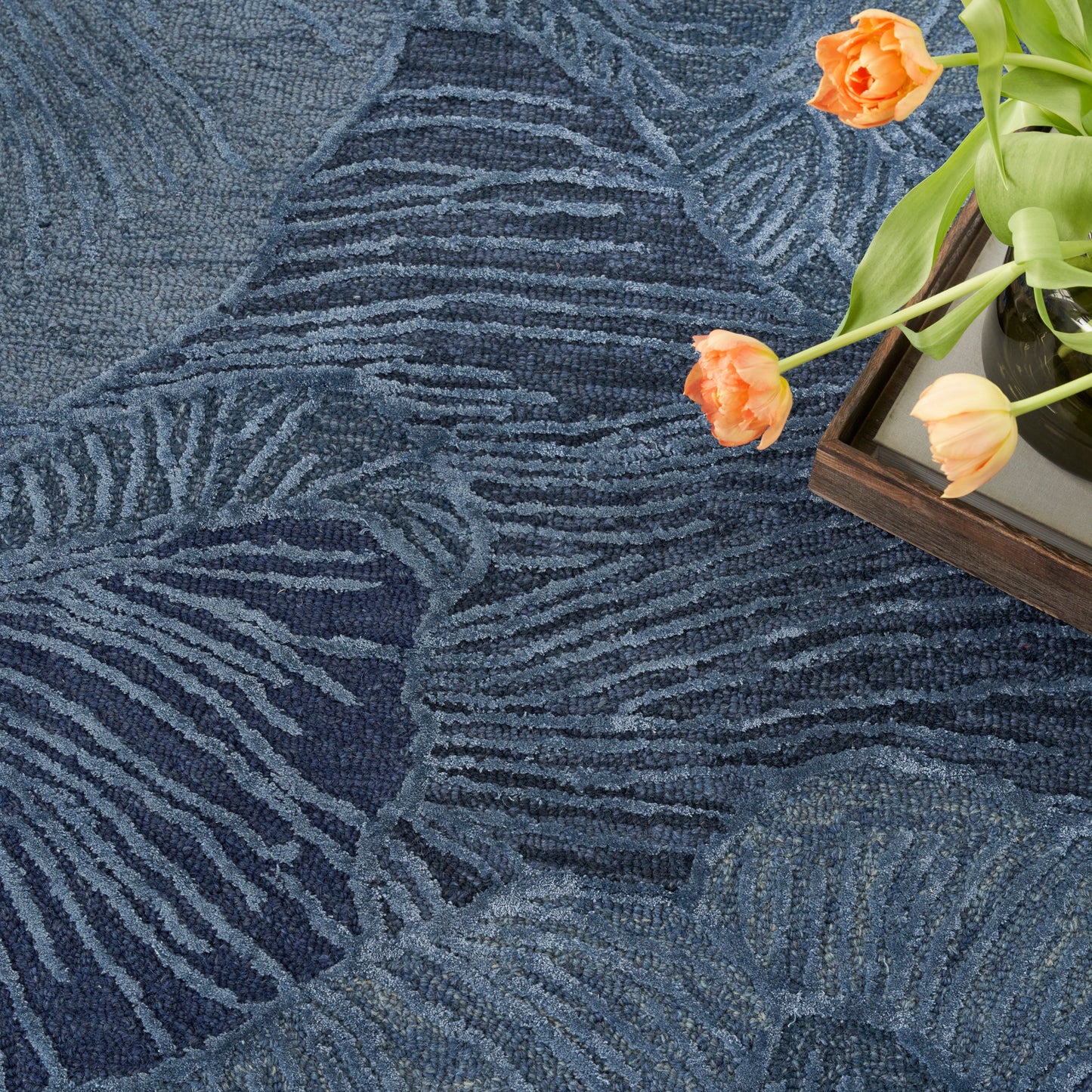 Dark blue textured rug with pale palm-leaf pattern; wooden tray with orange tulips resting on it.