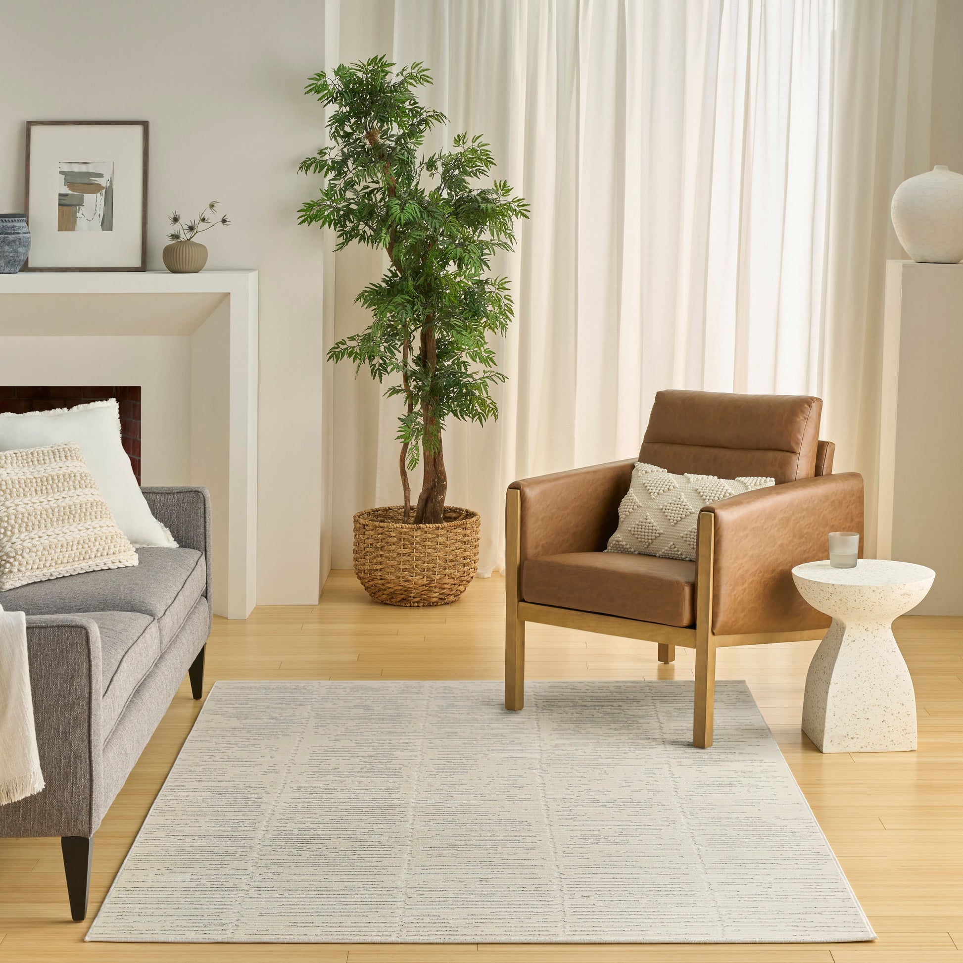 Modern living room with gray fabric sofa, brown leather armchair, light wood floor, beige geometric rug, and tall plant by white curtains.