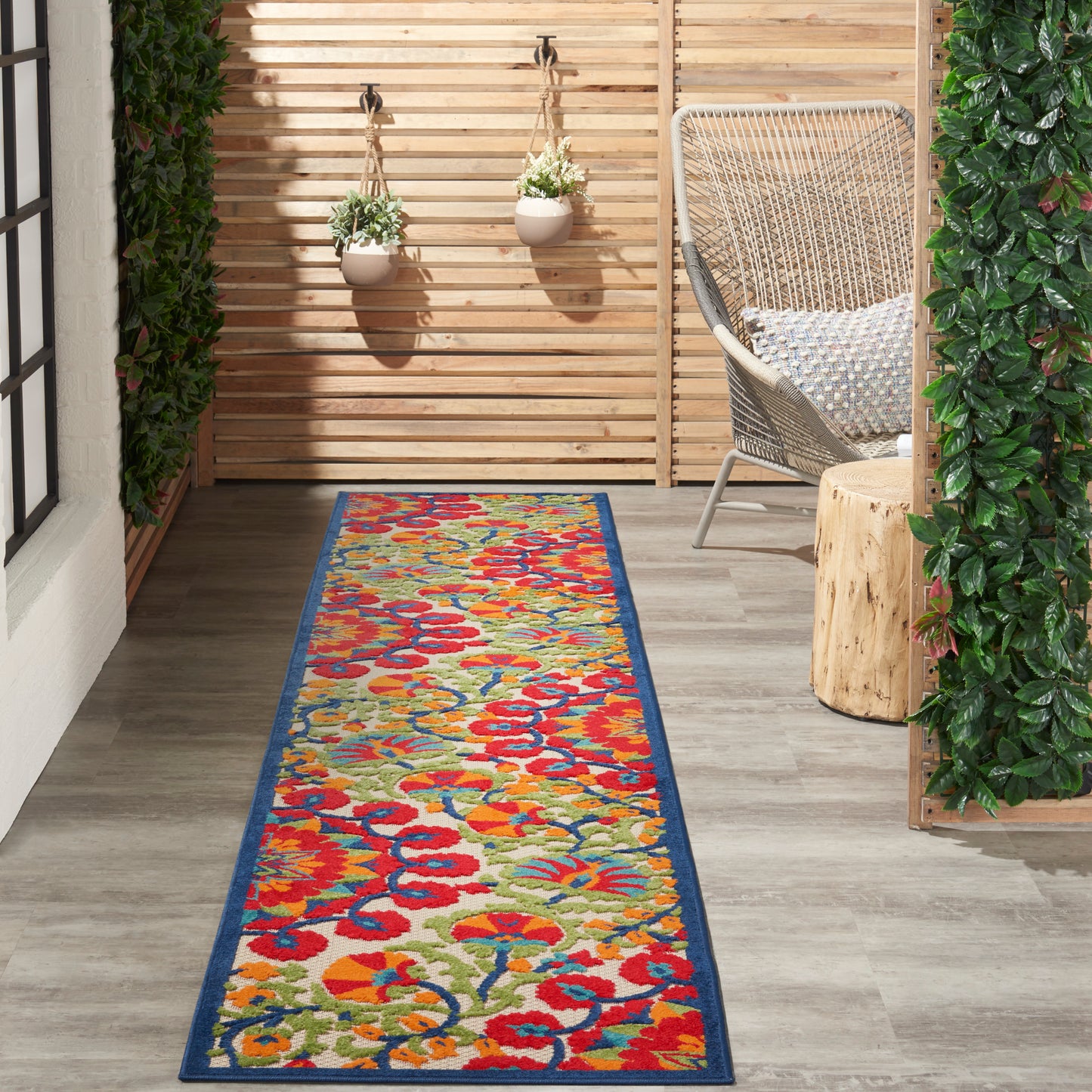 Colorful floral runner rug with blue border on a wooden patio beside a woven wicker chair and hanging planters.