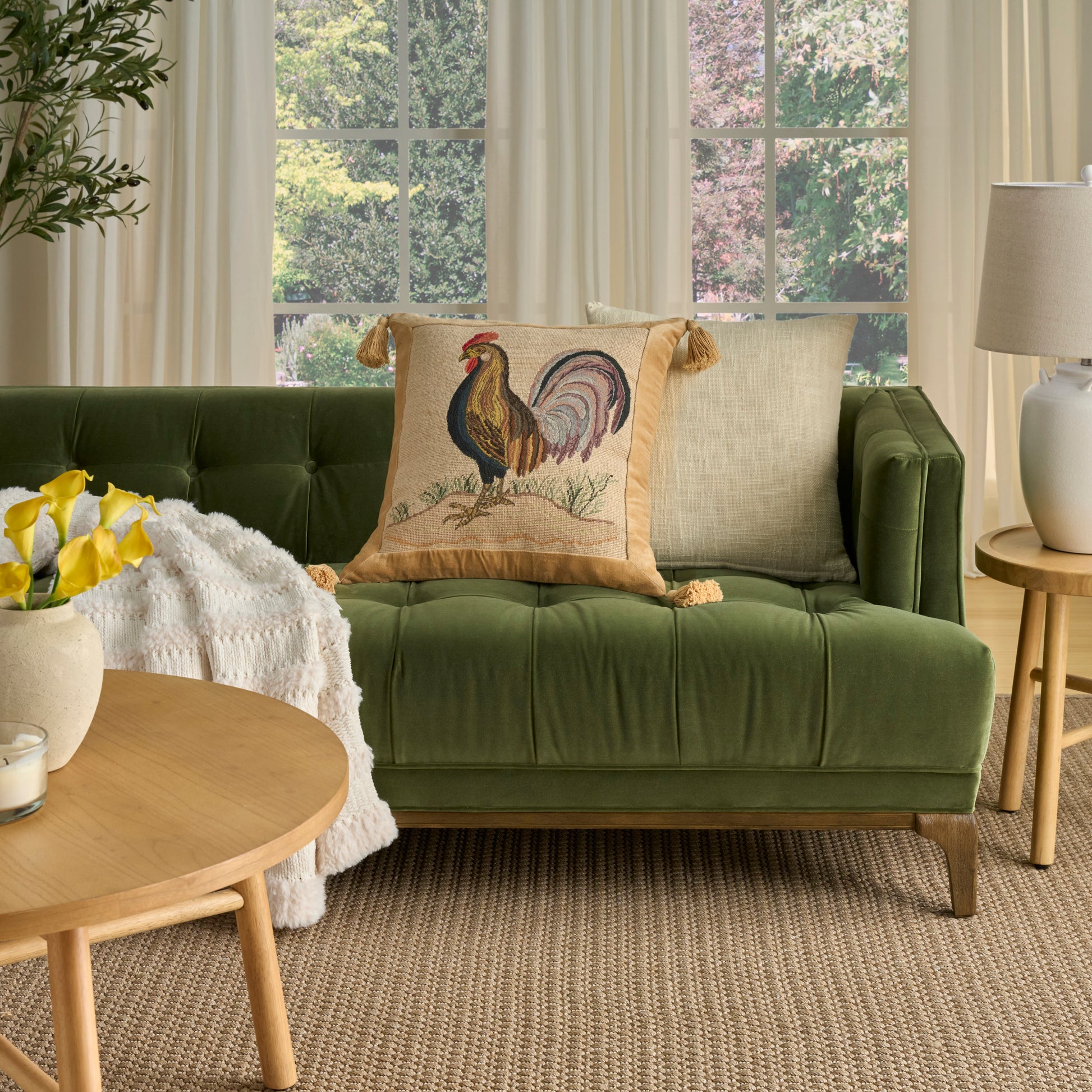 Green tufted sofa with rooster-embroidered pillow, beige pillow, round wood coffee table, yellow flowers, plant, lamp.