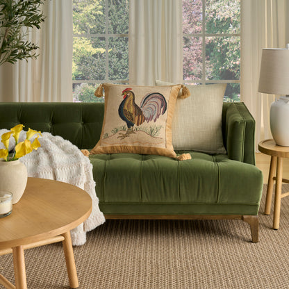 Green tufted sofa with rooster-embroidered pillow, beige pillow, round wood coffee table, yellow flowers, plant, lamp.