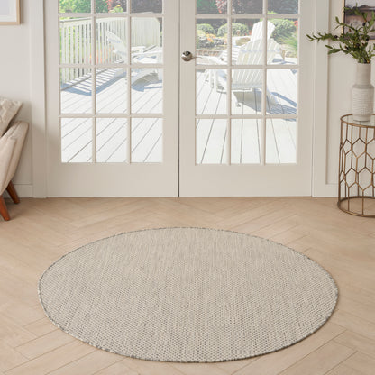 Round beige woven rug on light wood floor in front of glass French doors opening to an outdoor deck.