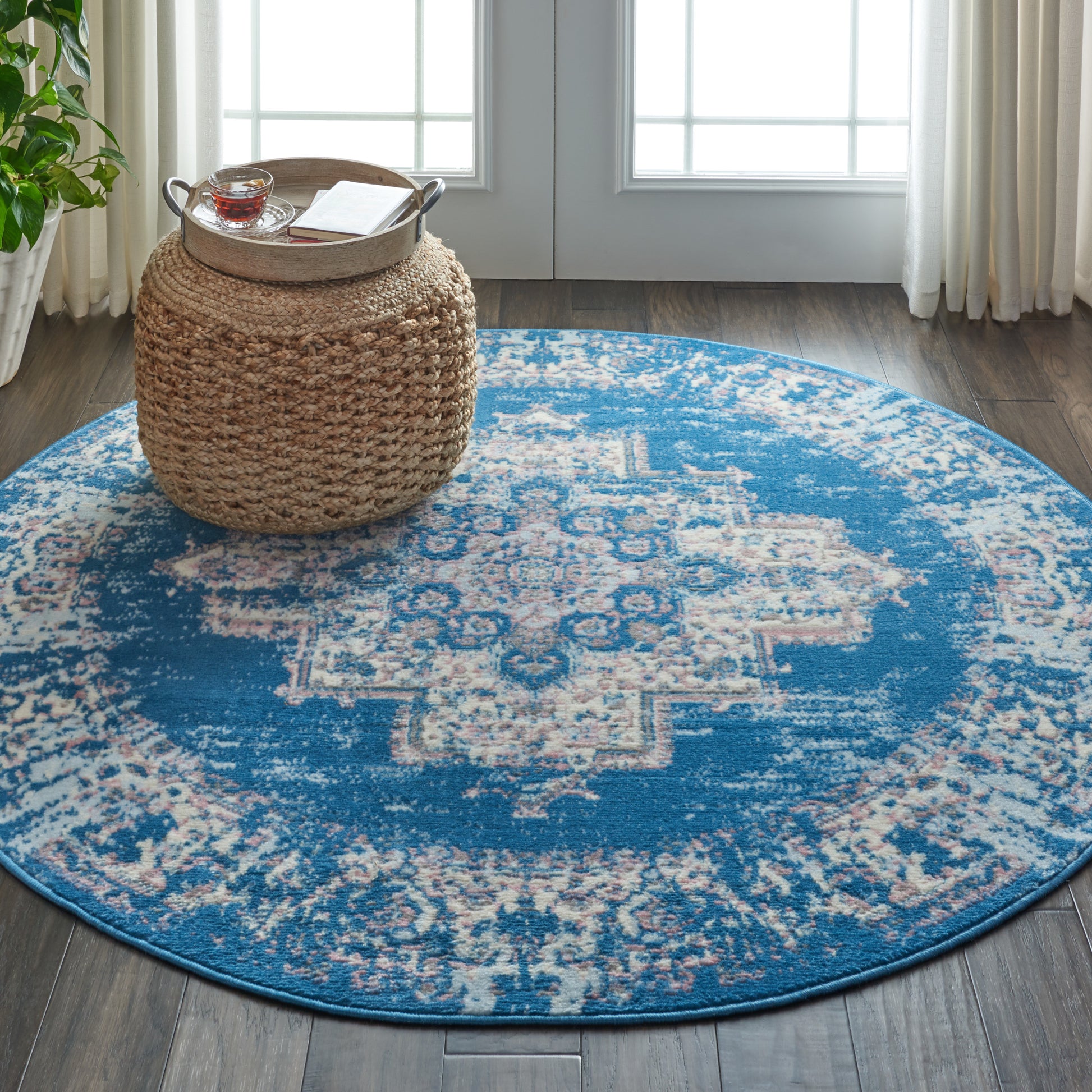 Round blue distressed area rug with ivory geometric center; natural woven ottoman with tray, tea, and a book.