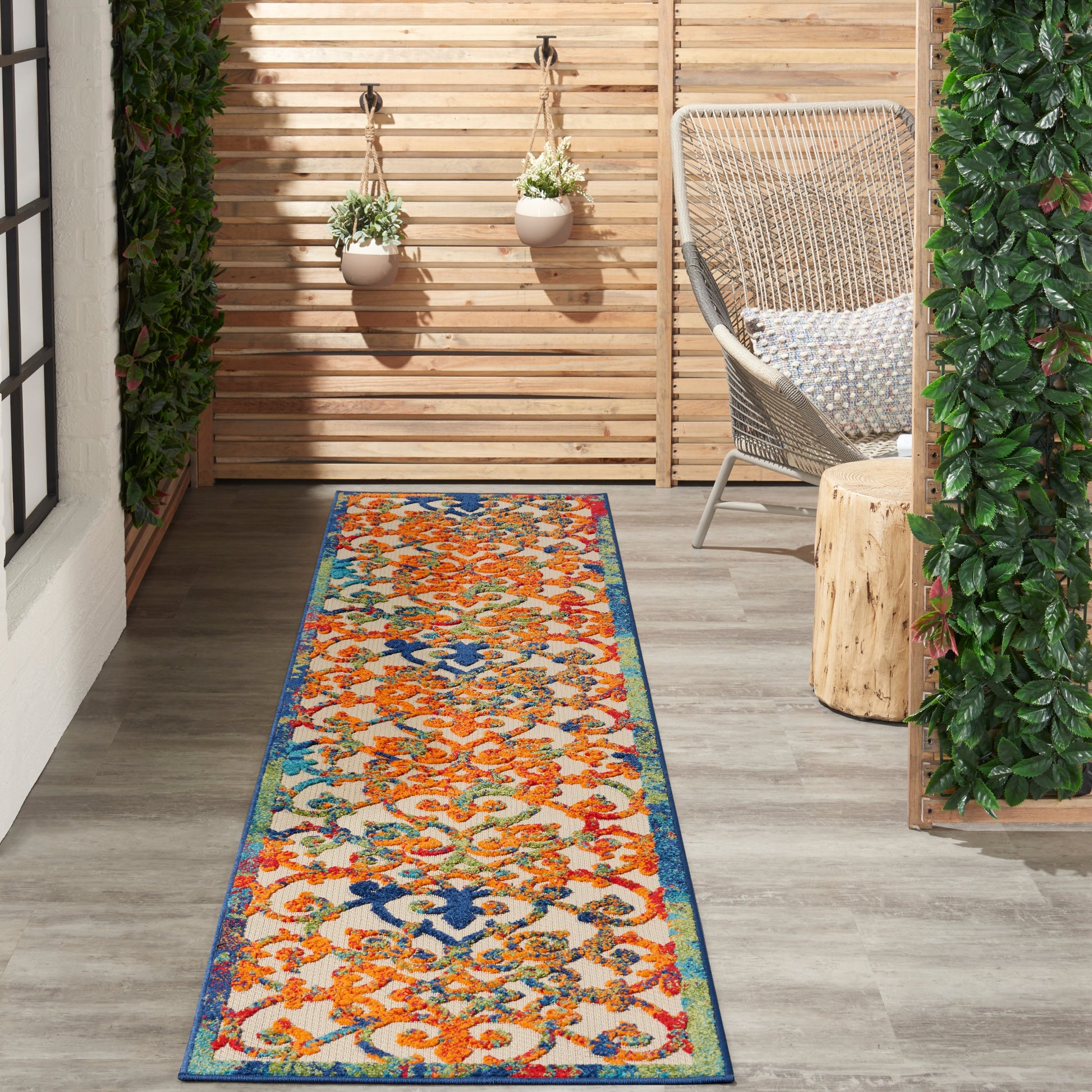 Outdoor patio runner rug with orange, blue, and green floral pattern on a wooden deck, wicker chair and hanging plants.